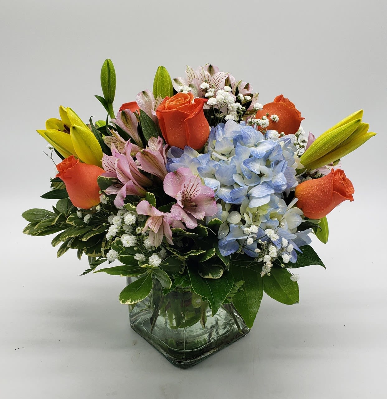 Mixed bouquet of orange roses, blue hydrangea, and yellow lilies in a glass vase
