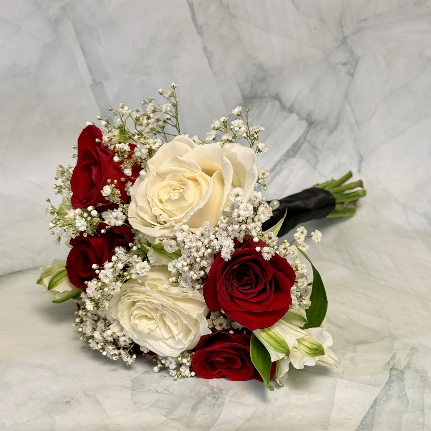 Red Rose and White Handheld - Deep, velvety red roses paired with creamy white peruvian lilies and delicate white filler blooms create a timeless handheld bouquet perfect for weddings, prom, or any romantic occasion. The red-and-white contrast reads classic and formal-she'll feel genuinely celebrated when presented with this elegant, compact arrangement.  This European hand-tied style bouquet includes roses, alstroemeria, and other white filler flowers arranged in a rounded, compact shape for rich texture and a soft, cloudlike edge. It's finished with a ribbon and a black stem wrap and is shown presented in a clear glass vase to keep the focus on the blooms. Ideal as an anniversary or wedding bouquet or as a prom accessory, it's sized to be comfortably carried and enjoyed fresh-snip stems and change water daily to extend vase life.