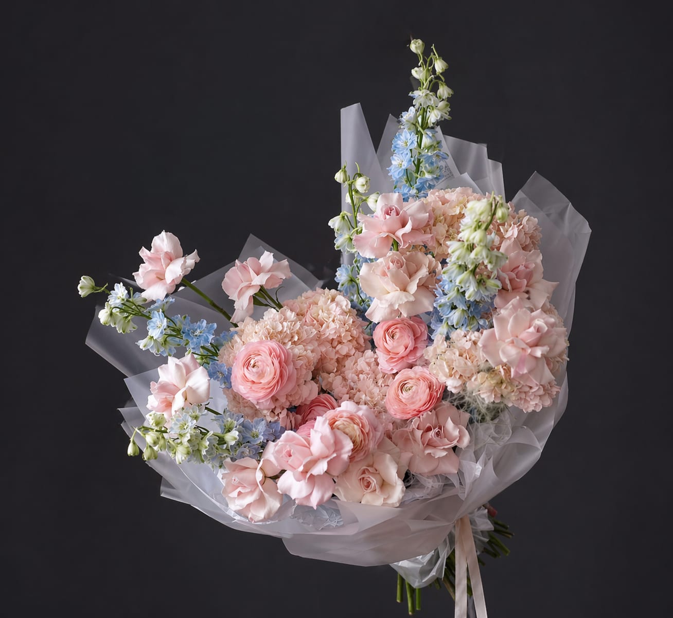 Blush Cloud Garden - Blush Cloud Garden is a luxurious floral composition designed in soft pastel tones of blush pink, cream, and delicate blue. This bouquet features premium hydrangea, ranunculus, garden roses, carnations, and airy accents of delphinium, carefully arranged to create a light, cloud-like silhouette. Hydrangea adds fullness and softness to the design, creating a rich base that enhances the layered texture of the bouquet. Ranunculus and garden roses bring elegance and refinement, while delicate blue delphinium adds a subtle contrast and movement. The composition is designed with a focus on natural flow, volume, and depth, giving the bouquet an effortless, airy look while maintaining a structured, premium feel. Wrapped in translucent high-end paper, it presents beautifully for both gifting and display. Perfect for birthdays, anniversaries, romantic gestures, or as a sophisticated gift for someone who appreciates refined floral artistry. Each bouquet is handcrafted and may vary slightly to ensure the freshest seasonal flowers and the highest quality.