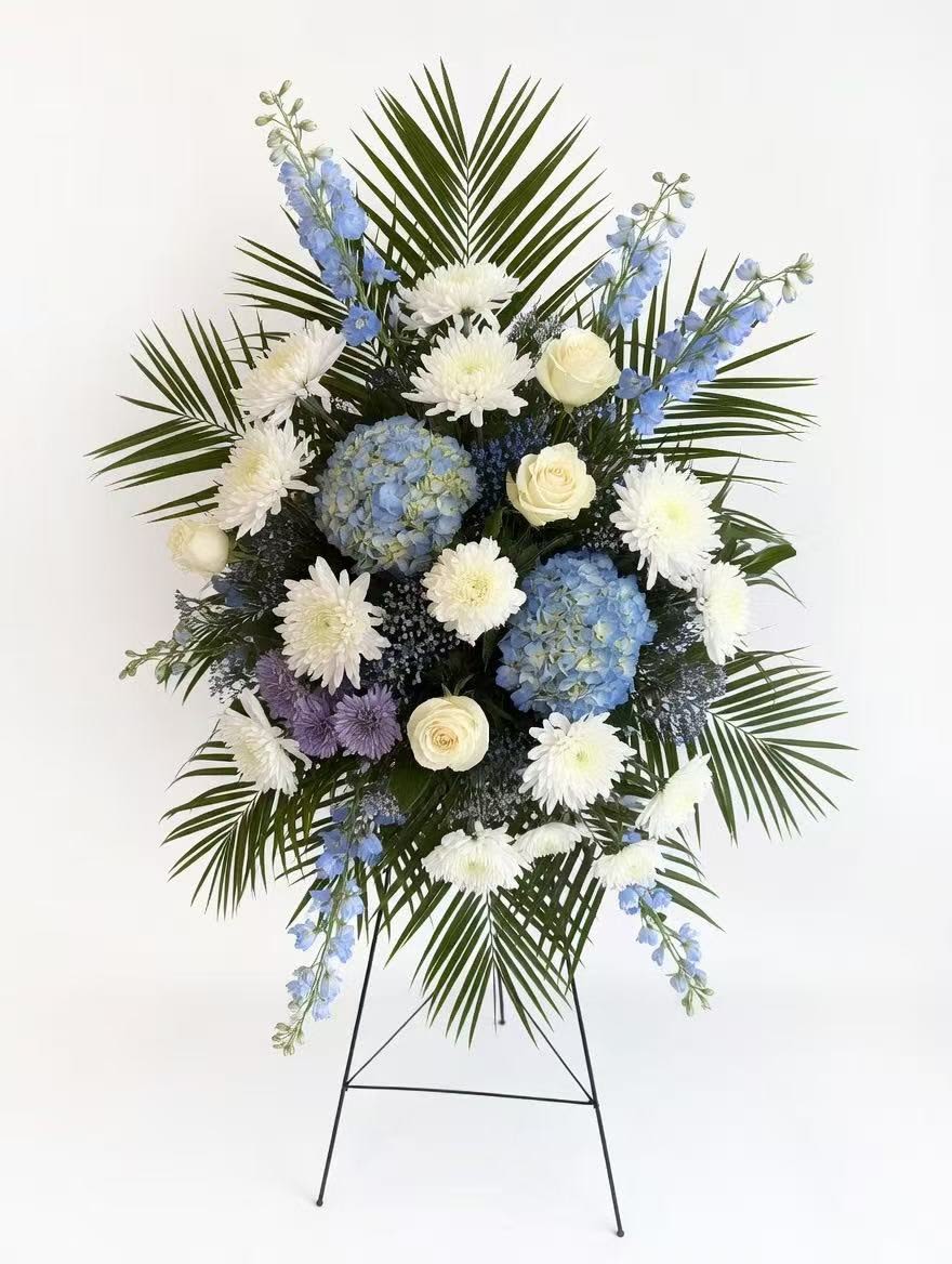 Sincere Thought Sympathy Spray with Blue Hydrangeas - A graceful standing sympathy spray that brings calm and reverence to a funeral or memorial service, this arrangement pairs blue hydrangeas with white chrysanthemums and cream roses for a serene, respectful display. The Sincere Thought Sympathy Spray with Blue Hydrangeas is designed for remembrance-its cool white, blue, lavender, and green palette quietly honors the person you're thinking of and helps convey heartfelt condolences.  Cream roses, pale blue hydrangeas, white chrysanthemums, and touches of delphinium are arranged in a large, symmetrical spray with long palm fronds radiating behind the flowers to create a formal, layered silhouette and strong vertical presence. Mounted on a metal easel for easy display at a service, the design reads traditional and ceremonial; the soft textures and cool colors evoke peaceful comfort.  Perfect for a funeral, memorial, or to send to a family home as an expression of sympathy, this spray offers a dignified way to say you care and to help loved ones feel supported. The standing format on the included metal easel makes delivery and placement simple-no extra hardware needed.