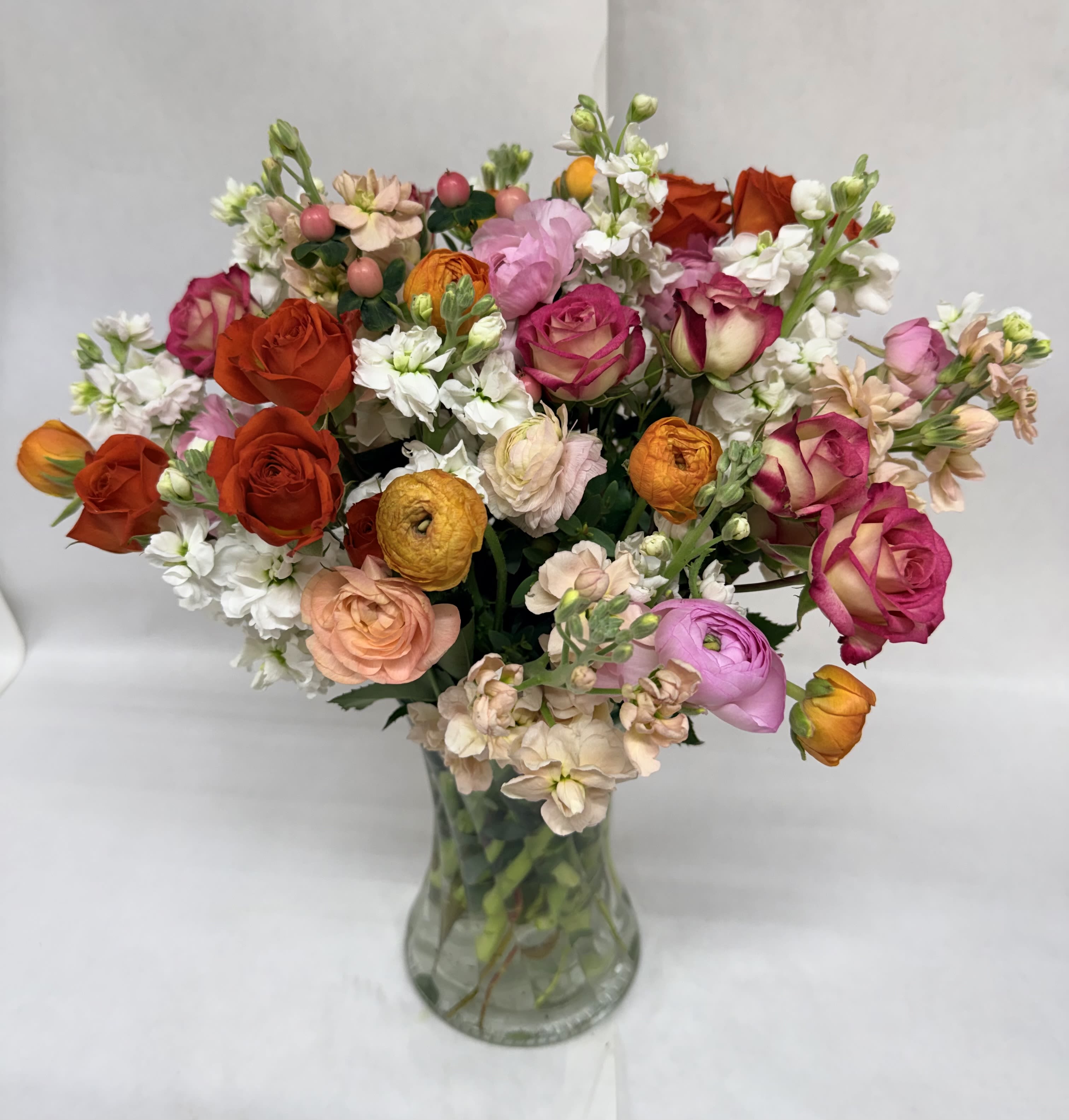 Citrus Linen Bouquet - A lively, garden-style cascade of pink, orange, peach and white sets the tone - the kind of birthday bouquet or anniversary flowers that makes her feel warmly celebrated. This Citrus Linen flower arrangement pairs lush roses and layered ranunculus with delicate stock and tiny white filler blooms to create a dense, rounded that's both bright and romantic for Mother's Day or any special occasion.  The flowers are arranged compactly in a clear glass vase for a classic, unfussy presentation that lets the colors pop. Textural contrasts between the velvety roses, the ruffled ranunculus, and the airy stock give the bouquet a full, tactile look while maintaining an elegant silhouette that suits a dining table or bedside. The palette reads fresh and uplifting - peach and orange notes add warmth, pink brings softness, and white keeps it light.  This bouquet is designed to make the recipient feel truly seen and celebrated - perfect for moms, partners, or friends marking another year or milestone. Arrives ready to display in its vase; keep in fresh water away from direct sunlight and change the water every other day to extend vase life.