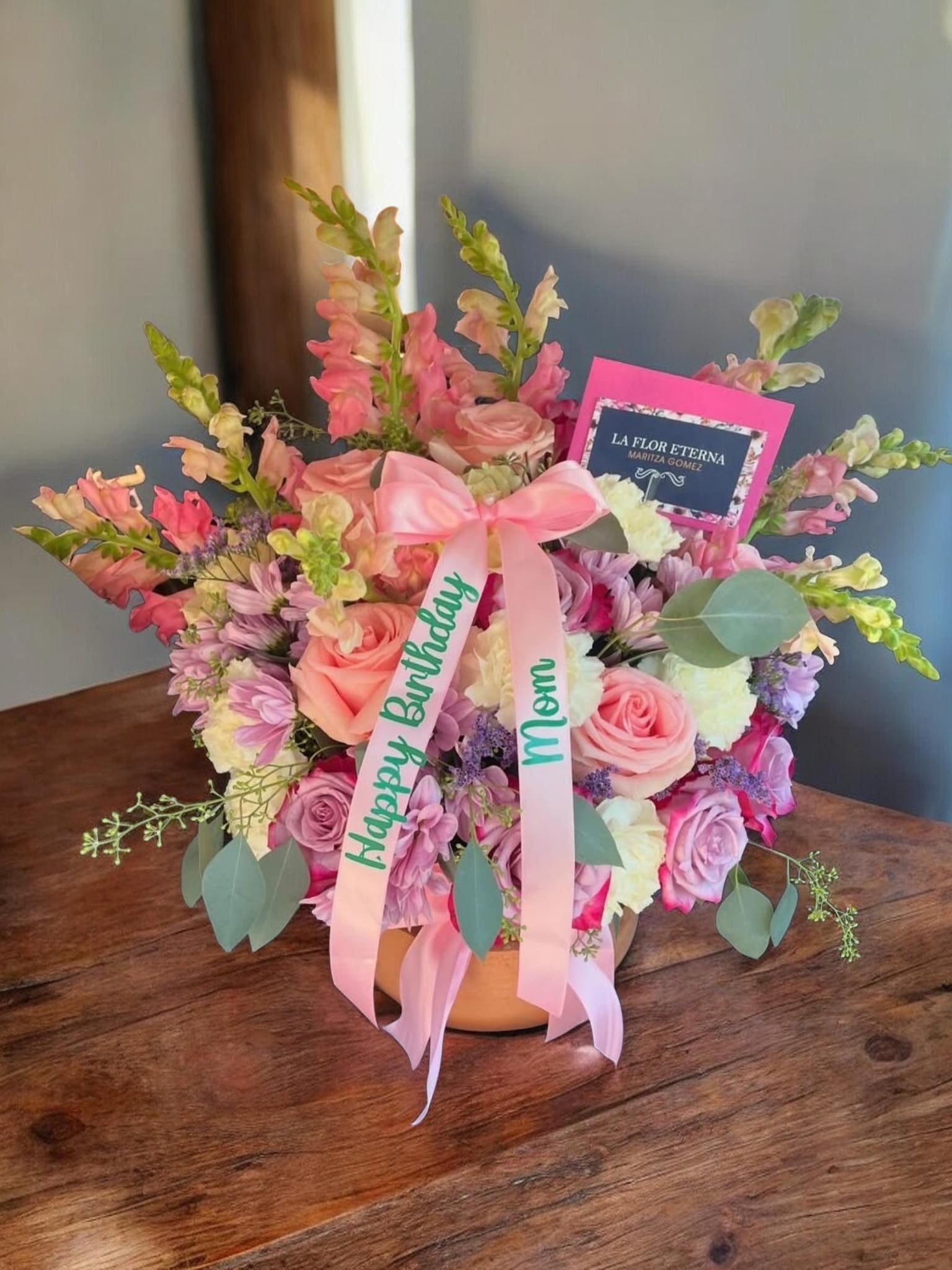 Ollita de Mama - A soft, rounded bouquet of pink and peach roses accented with tall snapdragons and lavender stock creates a gently romantic birthday bouquet that also feels right for Mother's Day, an anniversary, or a thank-you gift. The mix of daisies, white carnations and clustered hydrangea-like blooms adds playful texture while eucalyptus and airy green foliage soften the silhouette - the kind of arrangement that makes the person you're thinking of feel seen and celebrated.  Arranged in a painted or decorated round ceramic vase (or presented in a round hat box on some options), this design is finished with layered satin ribbons - typically a pink or lavender . Sizes are built to read lush and abundant: a dense main cluster with taller snapdragon or stock spikes rising above for height and movement. Colors run through pink, peach, white, purple and soft green for a pastel, feminine palette.  Choose this arrangement when you want a heartfelt, hand-delivered impression: it's ideal for personal celebrations where you want her to feel cherished. Orders come with the vase or box presentation and a gift card; for special requests (message on the card, preferred ribbon color, or specific vase option) please note them at checkout.