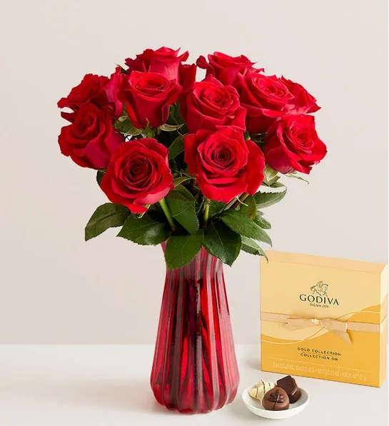 Red roses in a red glass vase with a box of chocolates beside them