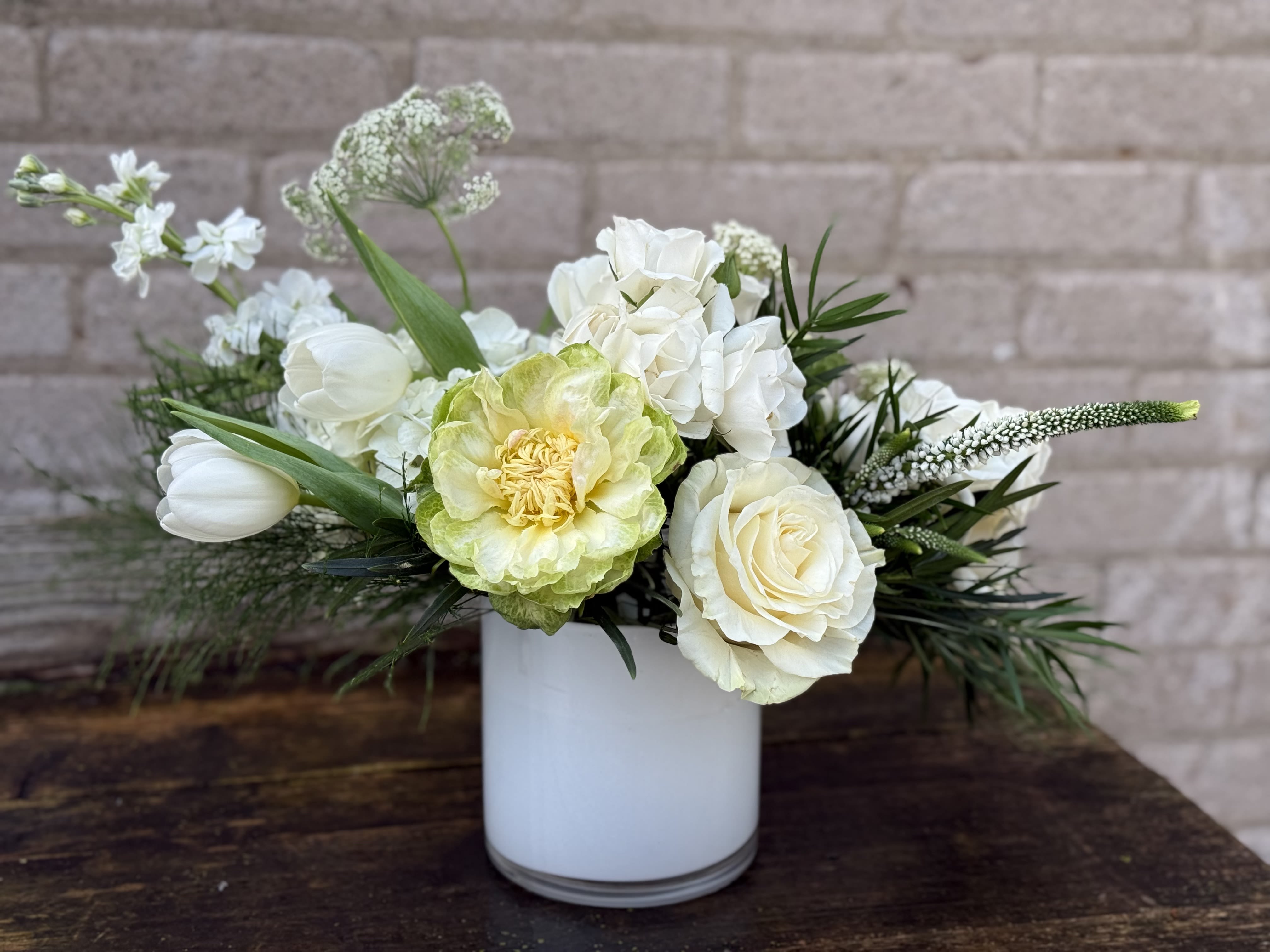 Fresh White - A serene, low-profile floral arrangement perfect for sympathy, a wedding table, or simply to brighten someone's day. Fresh White centers white roses and tulips around a pale green focal bloom, creating an understated yet elegant bouquet that reads calm and refined-an ideal choice when you want the person you're thinking of to feel seen and comforted.  The composition is horizontal and low, arranged in a clean white ceramic vase that keeps the presentation modern and gift-ready. Airy white filler flowers and spiky dark foliage extend gently outward, adding texture and depth against the soft palette of white, green, and a hint of yellow. This mix of roses, tulips, and complementary seasonal blooms creates a balanced, tactile display that looks lovely on a dining table, mantle, or bedside.  Designed to convey quiet elegance, Fresh White suits memorial moments, intimate celebrations, or everyday gestures of care. The compact low shape fits most surfaces; change water every two days and trim stems slightly to help blooms last. Orders include the white ceramic vase for immediate display.