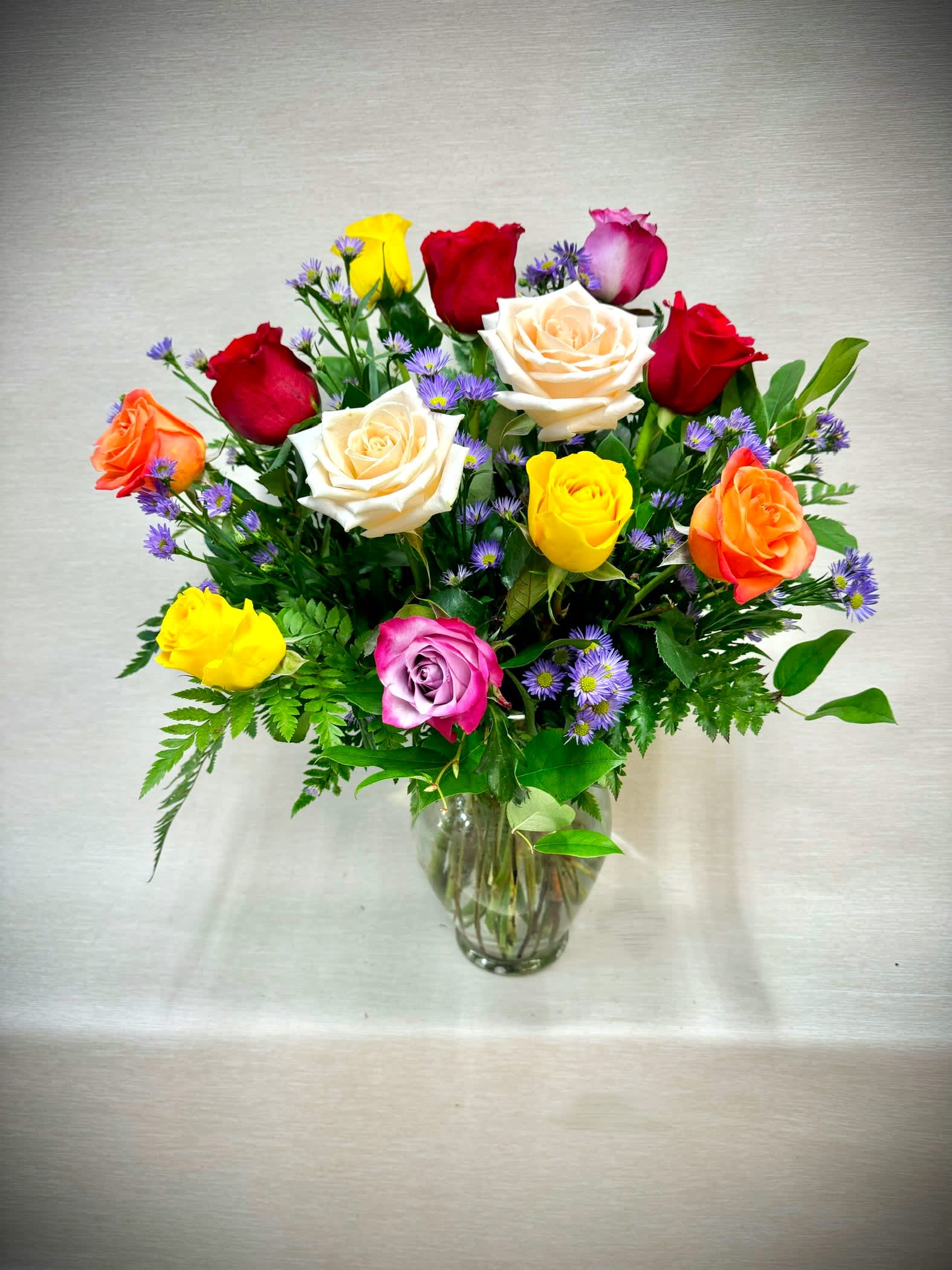 Mixed bouquet of multicolored roses and small purple flowers arranged in a clear glass vase