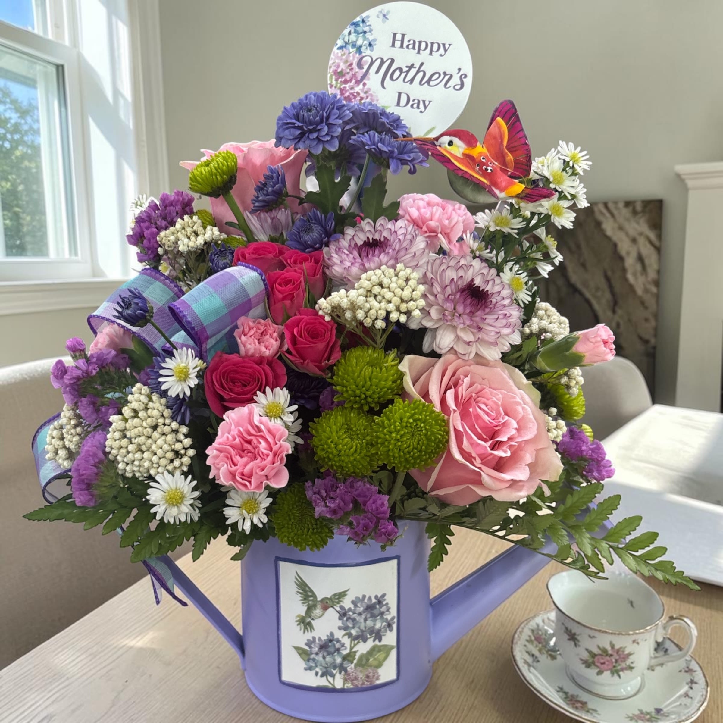 Fluttering Garden Watering Can  - Celebrate Mom with our exclusive Fluttering Garden Watering Can from Aspen Tower Florist in Greensburg, Indiana. This cheerful Mother’s Day flower arrangement is designed in a reusable metal watering can available in lavender or periwinkle and filled with a fresh mix of pink, purple, white, and green seasonal flowers. Accented with a decorative hummingbird pick and festive Mother’s Day sentiment, it’s a gift that feels joyful, thoughtful, and ready to display.  Perfect for Mother’s Day delivery to homes, offices, nursing homes, assisted living communities, and care facilities, this design brings spring garden charm indoors. After the flowers are enjoyed, the keepsake watering can can be reused for plants, décor, or gardening.  Customers love this arrangement for its reusable container, fresh seasonal blooms, soft spring colors, and one-of-a-kind garden styling. It is handcrafted and hand-delivered by Aspen Tower Florist, a real local florist proudly serving Greensburg, Indiana and surrounding areas including Batesville, Brookville, Rushville, Shelbyville, Westport, Hope, North Vernon, Columbus, and nearby communities.  Flower varieties and shades may vary based on freshness and availability while maintaining the overall style, color palette, and value. Order early for Mother’s Day flower delivery in Greensburg, Indiana.