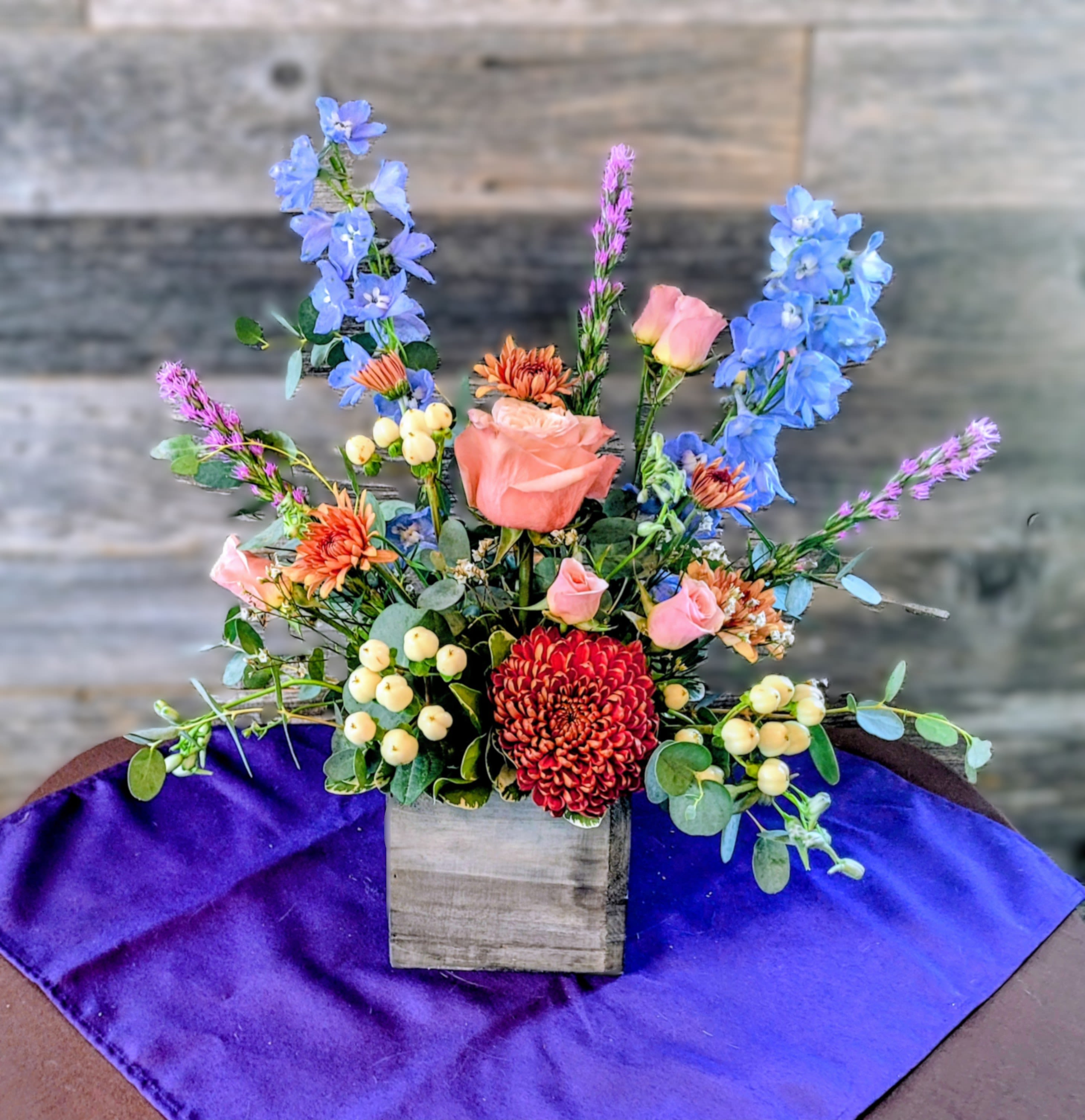 Mixed arrangement of pink roses, blue delphinium, and red mum in a wooden cube vase