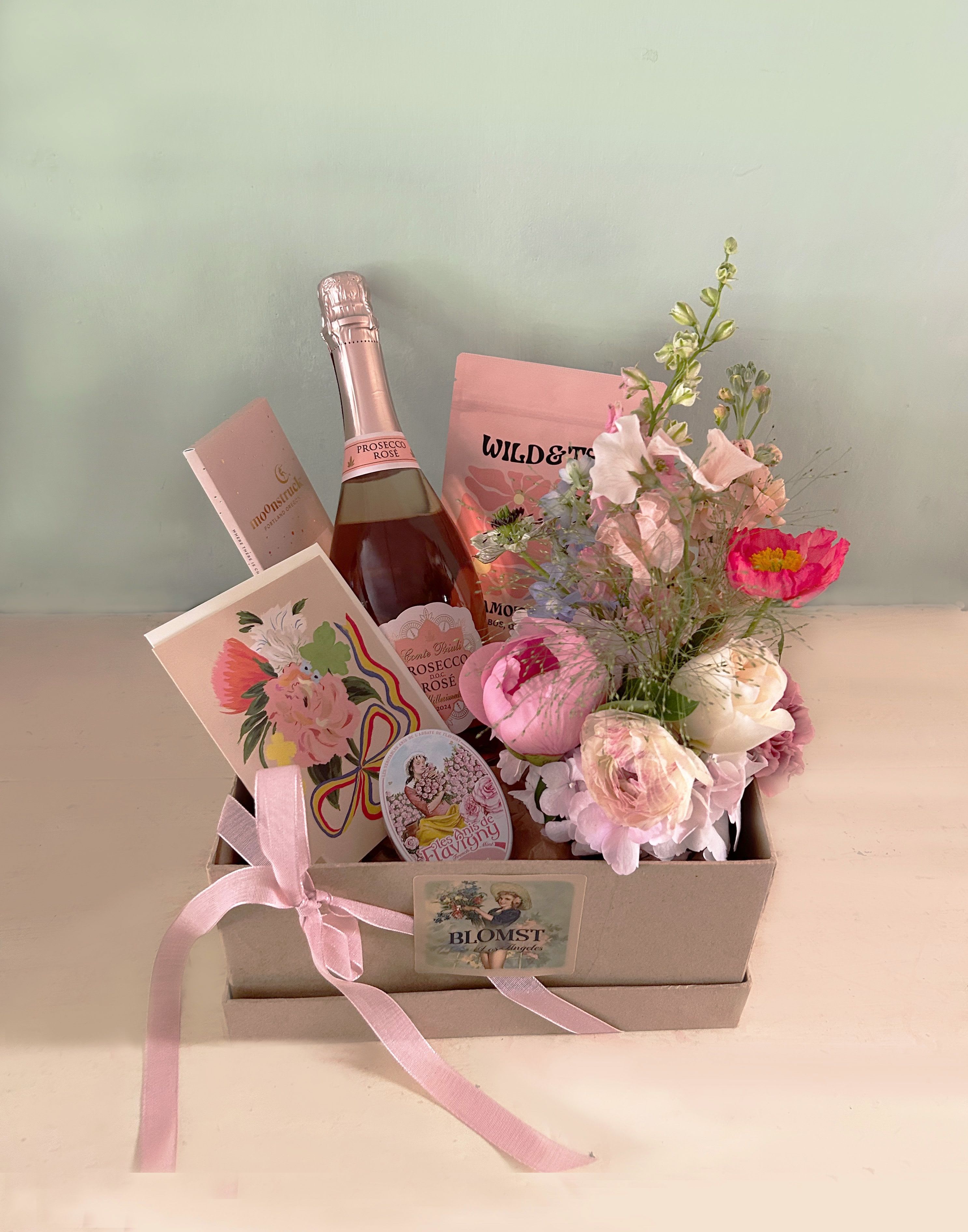 Beverly Bubbly Box - A celebration box with bubbly, chocolate, tea, rose mints, fresh flowers and a pretty card for your personal message.  Sparkling Prosecco Rosé wine that is made from both Glera (Prosecco) and Pinot Noir grapes. Wild &amp; Tea Bergamot Blossoms Herbal Tea. Luscious fruit with delicate floral undertones. Abundant in antioxidants to promote healthy, radiant skin. Organic Ingredients- Red rooibos, chamomile, orange peel, rosehips, cornflower, rose and bergamot oil. Moonstruck Creamy Milk Chocolate Bar, 68% Cacao. Handcrafted superior chocolate from Portland, Orgeon. Les Anis De Flavigny: Hard candies with a subtly minty taste. These all-natural candies are housed in a vintage decorative tin, with about 30 mints per tin. Each anise seed is coated in naturally-flavored sugar to produce round and smooth rose flavored mints that will delight the palate and senses. Beautiful printed greeting card with a bouquet/w envelope for your perosnal message. Adorned with a mini Blomst LA bouquet in a small cylinder vase  All placed in a reusable paper mache box with lid, tied with riboon, the box measures 10" by 10" by 5" Ready for hand delivery in LA