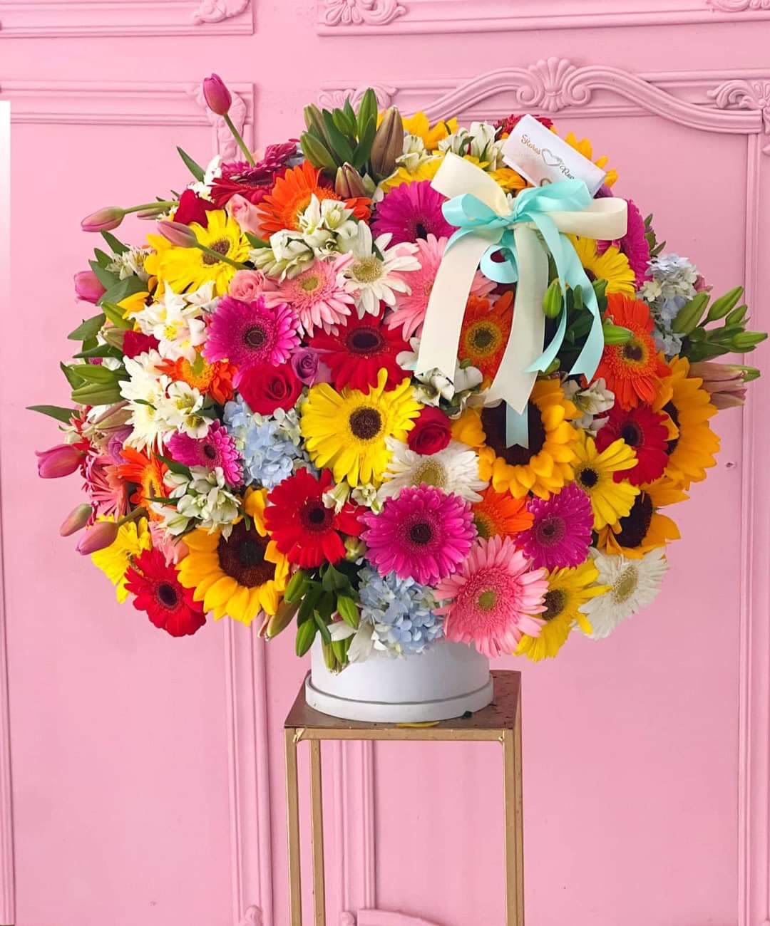 Vibrant Bloom Floral Box” - This floral box is a vibrant burst of color, a lively tribute for Mother’s Day. Inside the box, you’ll find exuberant gerberas, sunflowers, lilies, alstroemerias, and sky-blue hydrangeas, all blending with roses in a symphony of color. It’s perfect to surprise your wife this upcoming Mother’s Day, with a gift full of love and life.( The box is subject to changes and availability. We have it available in white, black, pink, and lilac.)