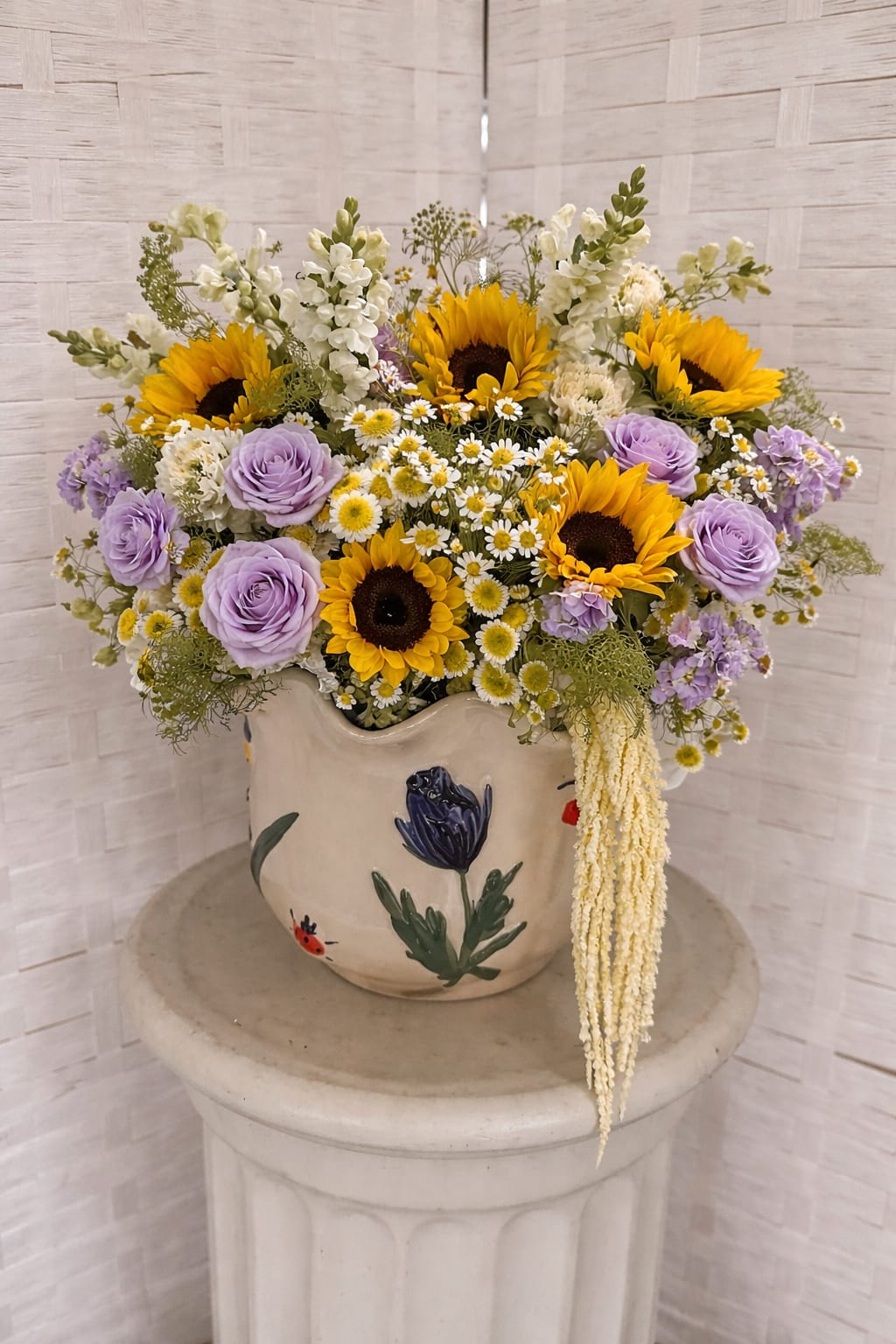 Serenity Garden for Mom - This radiant arrangement is a vibrant tribute for Mother’s Day. The sunflowers, in a warm yellow, are combined with lavender roses, white daisies, chamomile, and white snapdragons. All of this is presented in an exquisite hand-painted ceramic vase. This decorative base, with delicate details, becomes an ideal piece to honor our moms on this Mother’s Day and a decorative keepsake for the home that will last.