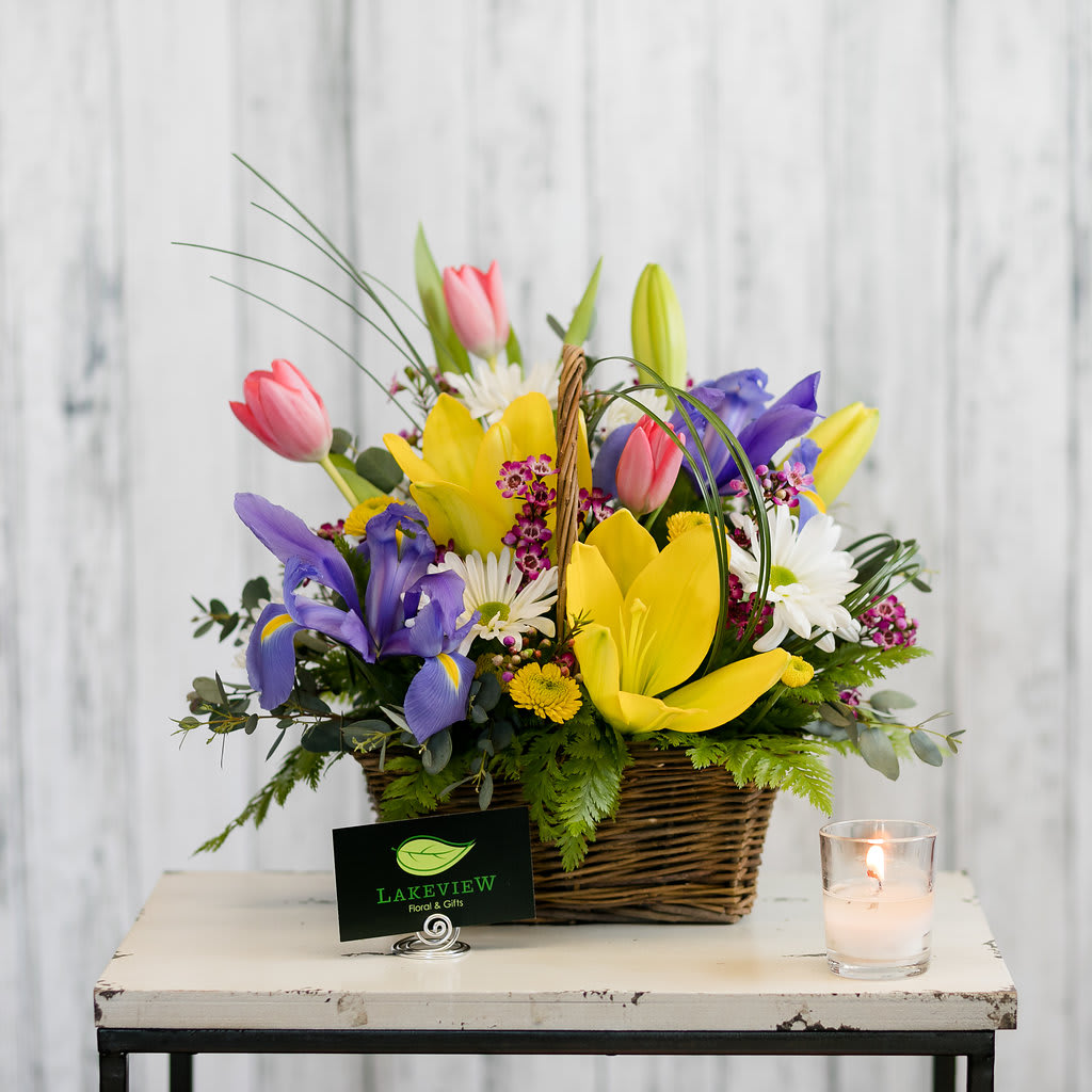 Basket arrangement of tulips, irises, and daisies with a candle beside it