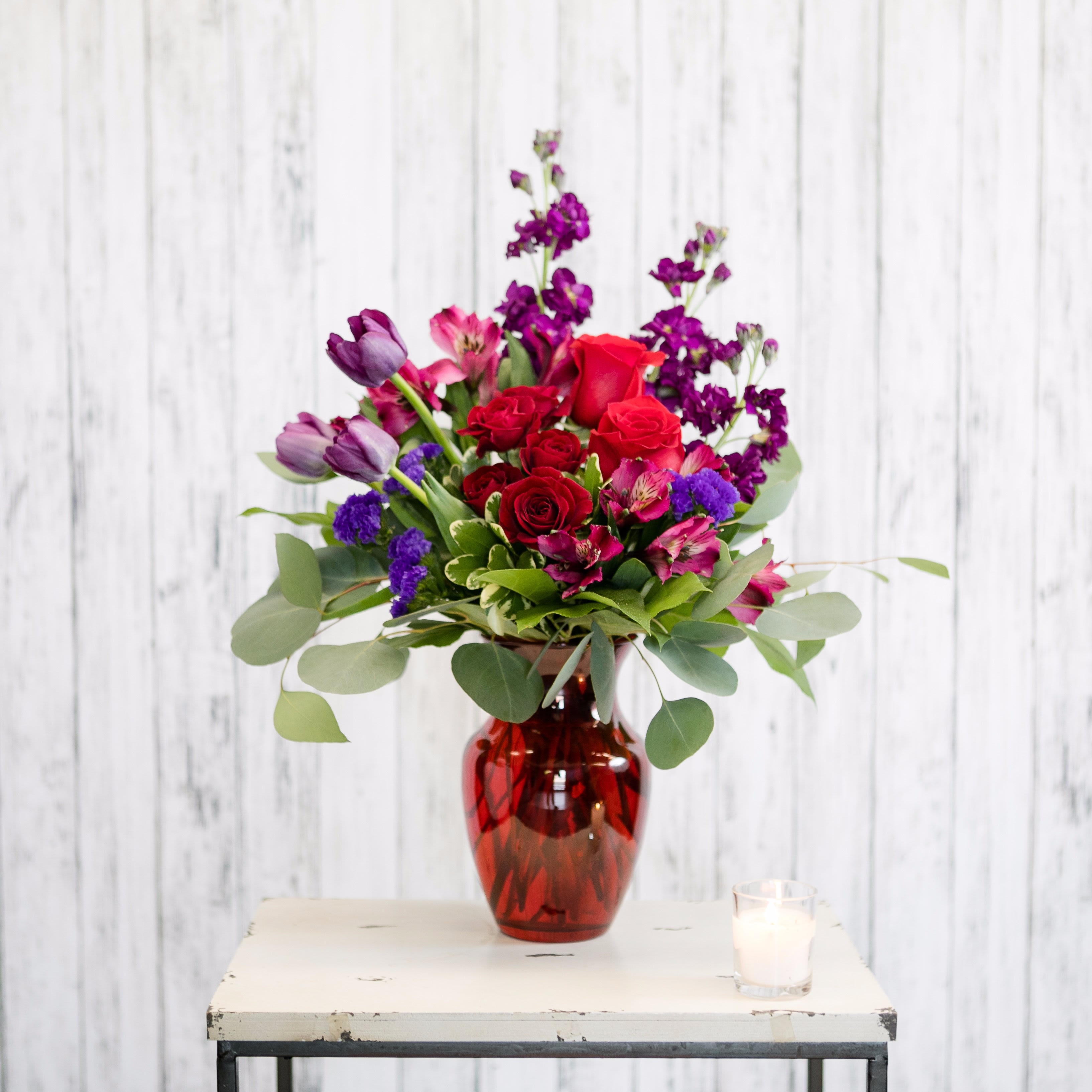 Red, pink, and purple mixed flower arrangement in a red glass vase on a small table with a candle
