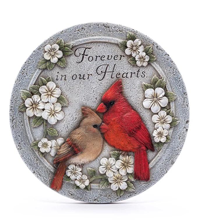 Forever in our hearts stone - A gently weathered stone plaque titled "Forever in our Hearts" offers a quiet, lasting tribute for sympathy or remembrance. This memorial arrangement piece places two raised birds - one bright red and one tan - amid delicate white blossom accents and soft pink and green touches, creating a wreath-like frame that reads as both comforting and hopeful for someone honoring a lost loved one.  The round decorative plaque has a stone-like finish and centered lettering that lends it weight and permanence. Small floral details wrap the birds, adding a soft contrast of color without overpowering the sentiment; the palette of white, red, pink and green keeps the look restrained and uplifting. Presented as a keepsake rather than perishable flowers, it's designed to sit on a mantel, grave marker area, or memorial table as a lasting expression of care.  Choose this piece when you want the person you're honoring to feel remembered and cherished-an understated way to say sympathy, remembrance, or condolences that lasts beyond fresh blooms. Ideal for sending to a family home, funeral service, or visiting a gravesite, the compact round size makes placement flexible and the durable finish ensures it endures as a tender reminder.