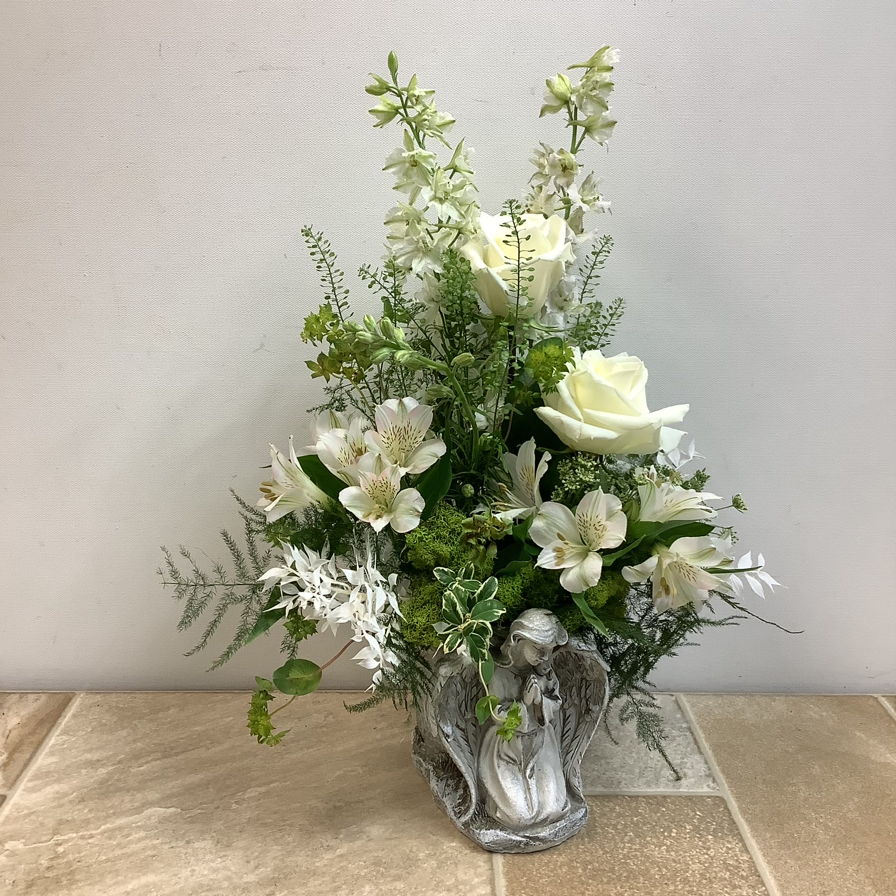 Enduring Beauty - This memorable floral arrangement, "Enduring Beauty" is an elegant and serene composition featuring a soft palette of white and green in a resin angel planter, making it especially fitting for sympathy or memorial occasions. The delicate white flowers include roses, alstroemeria, and larkspur. Accents may include ruscus, reindeer moss, and assorted greenery.   Approximately size: 14” wide x 17” tall  Primary flowers: rose, lily