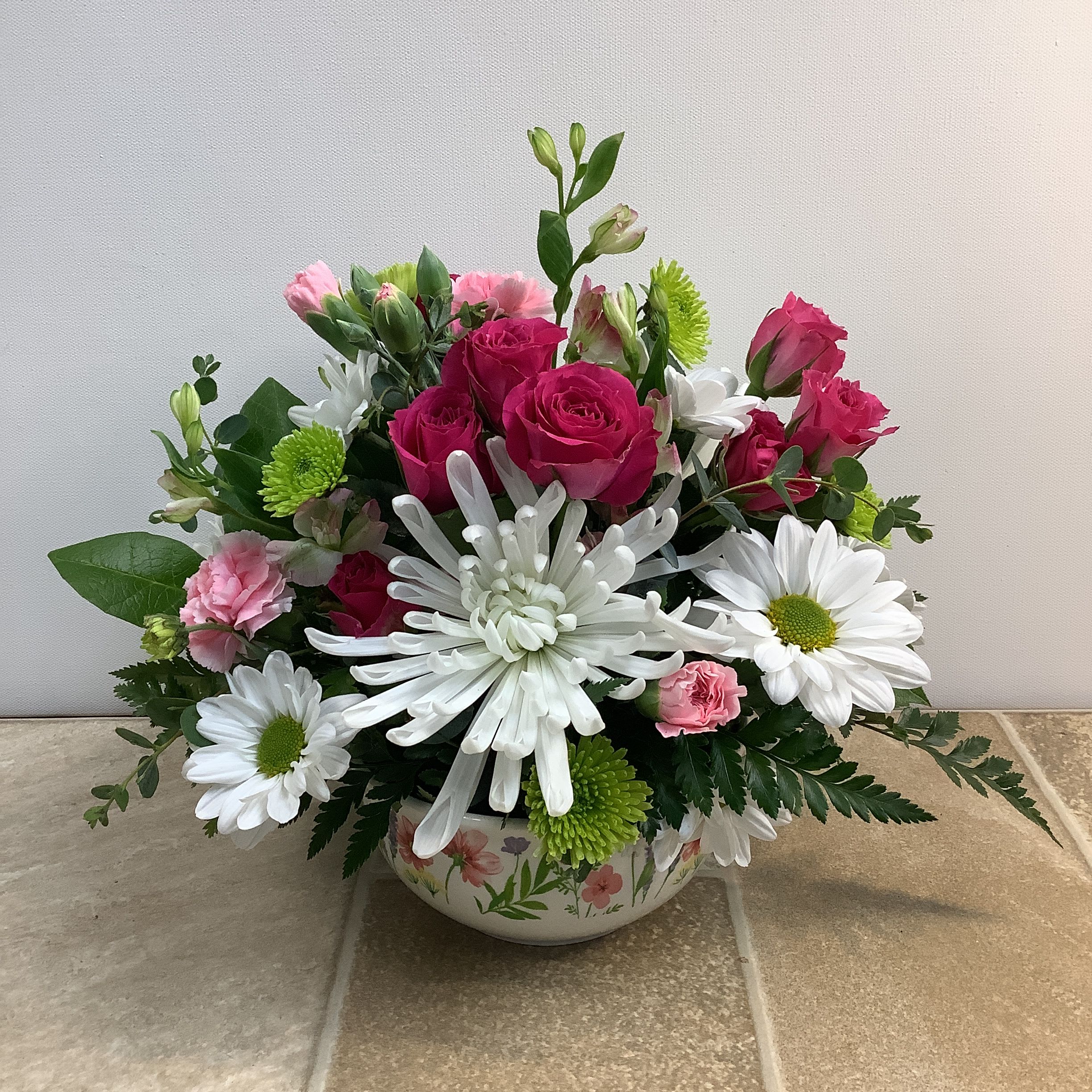 A Gentle Touch - “A Gentle Touch" features a delightful mix of blooms in a round, ceramic dish with a floral design. Dark pink spray roses and a white spider mum are the focal points. White daisies and green button mums add a fun, cheerful feel; and pale pink mini carnations and pink charmellia contribute a soft, romantic tone. A mix of lush greenery frames the arrangement beautifully  It measures approximately 12” wide x 10” tall.  Primary flowers: Daisy, mum, rose, carnation