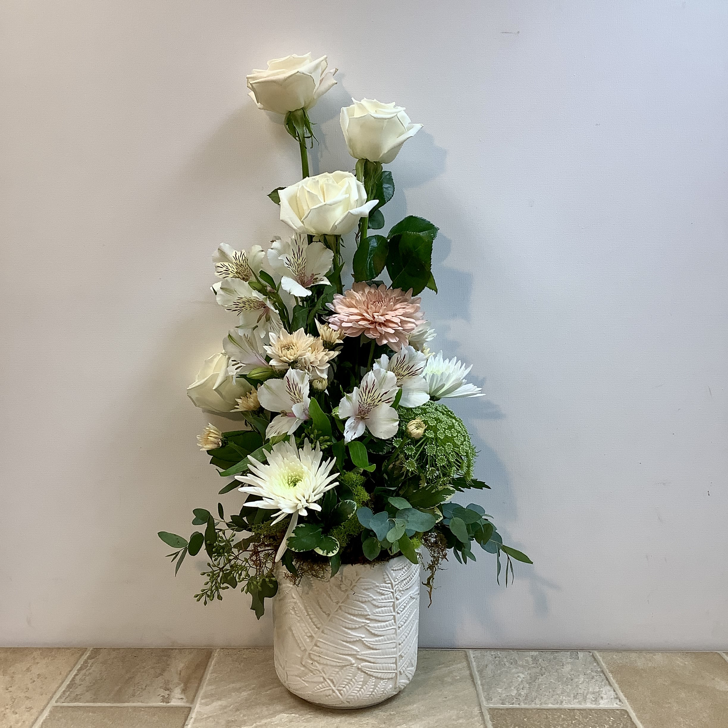 Chantilly Lace - This bouquet has a graceful, garden-inspired shape with soft neutral tones and fresh greenery.  The elegant, airy arrangement features creamy white roses rising above a lush garden mix of blooms. Soft white cremones, delicate alstroemeria, and a blush chrysanthemum create a gentle blend of textures and color. Accents of Queen Anne’s lace and fresh eucalyptus add a natural, woodland feel, while the flowers are arranged in a textured white ceramic container with a subtle botanical pattern. The design has a light, vertical silhouette that feels both modern and timeless—perfect for bringing calm beauty and natural charm into any space. Approximate size: 14” wide x 26” tall.  Primary flowers: rose, lily, mum