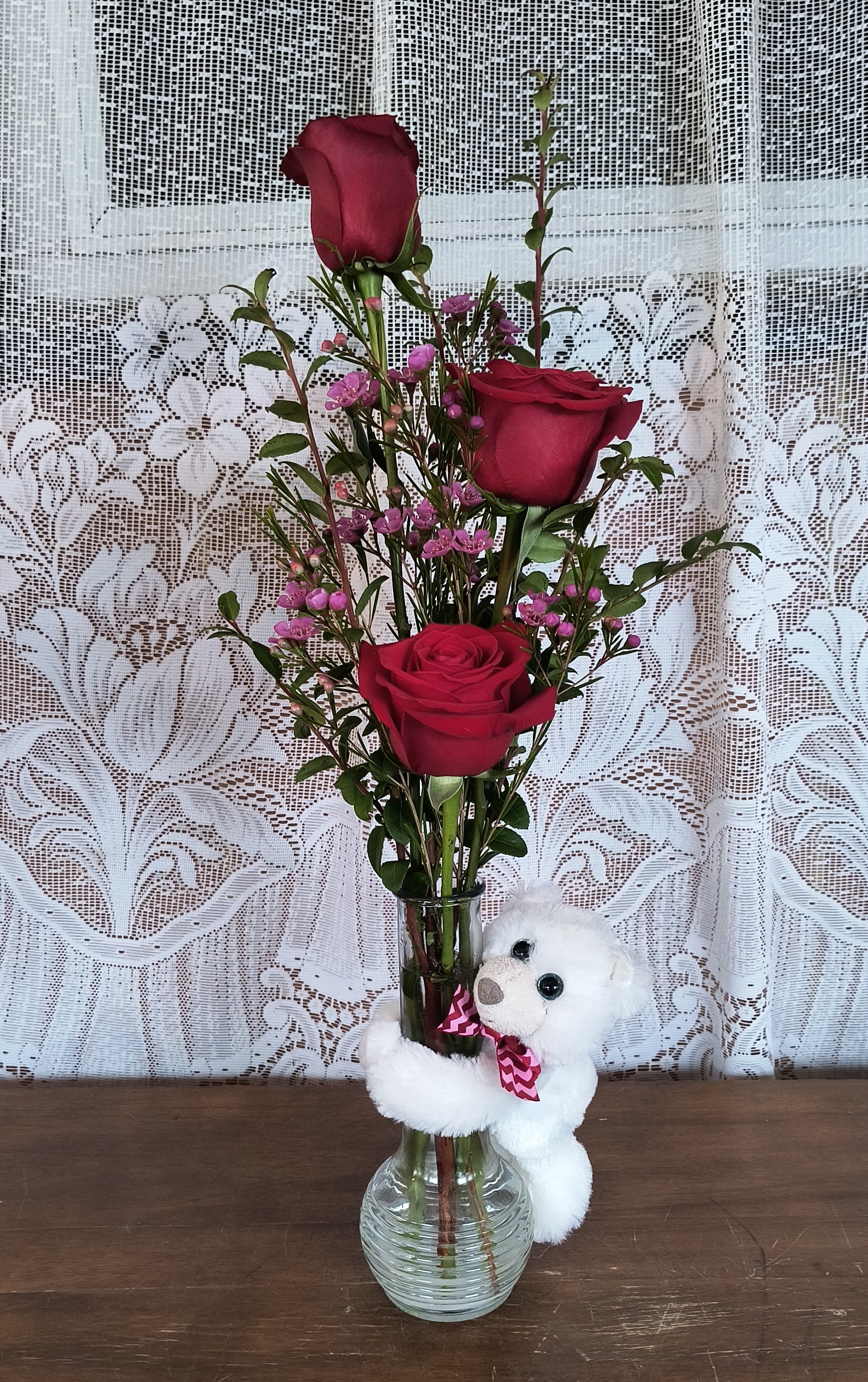 Three red roses in a glass vase with a small white teddy bear