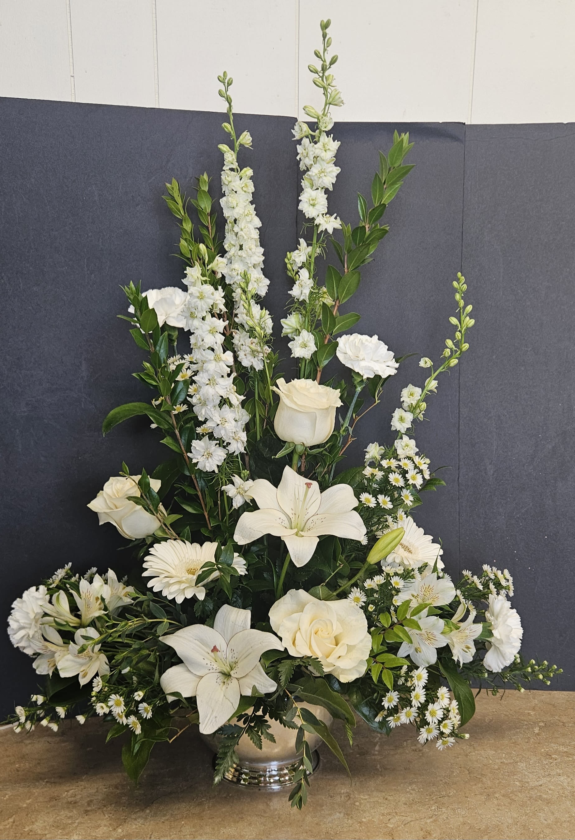 Peaceful White - A serene cluster of white flowers arranged around a devotional Madonna makes this sympathy arrangement a quiet, comforting choice for a funeral or memorial. The Peaceful White flower arrangement is designed as a sympathy centerpiece that reads as both reverent and gently uplifting - the person you're honoring will be surrounded by a calm palette of white and soft green that conveys sympathy and spiritual comfort.  White snapdragons and vertical sprays provide graceful height behind full white roses, open lilies, chrysanthemums, and daisy-like blooms, while baby's breath fills the composition for a soft, airy texture. Depending on the presentation you choose, the arrangement is offered in a low black tray with a small glass candle holder at the front for a votive, or as a taller formal display in a polished silver vase; both versions include the ceramic Madonna statue as the focal point and are arranged to sit respectfully on a memorial table or at a service.  Made to comfort the person you're thinking of, this piece is appropriate for services, home remembrances, or graveside gatherings - it announces your sympathy without overwhelming the moment. If selecting a tray, note the low-profile design suits tabletops and altars; the vase option gives added height for a display podium. Simple care: keep flowers in a cool place out of direct sun and ensure the water is refreshed every couple of days to maintain freshness.