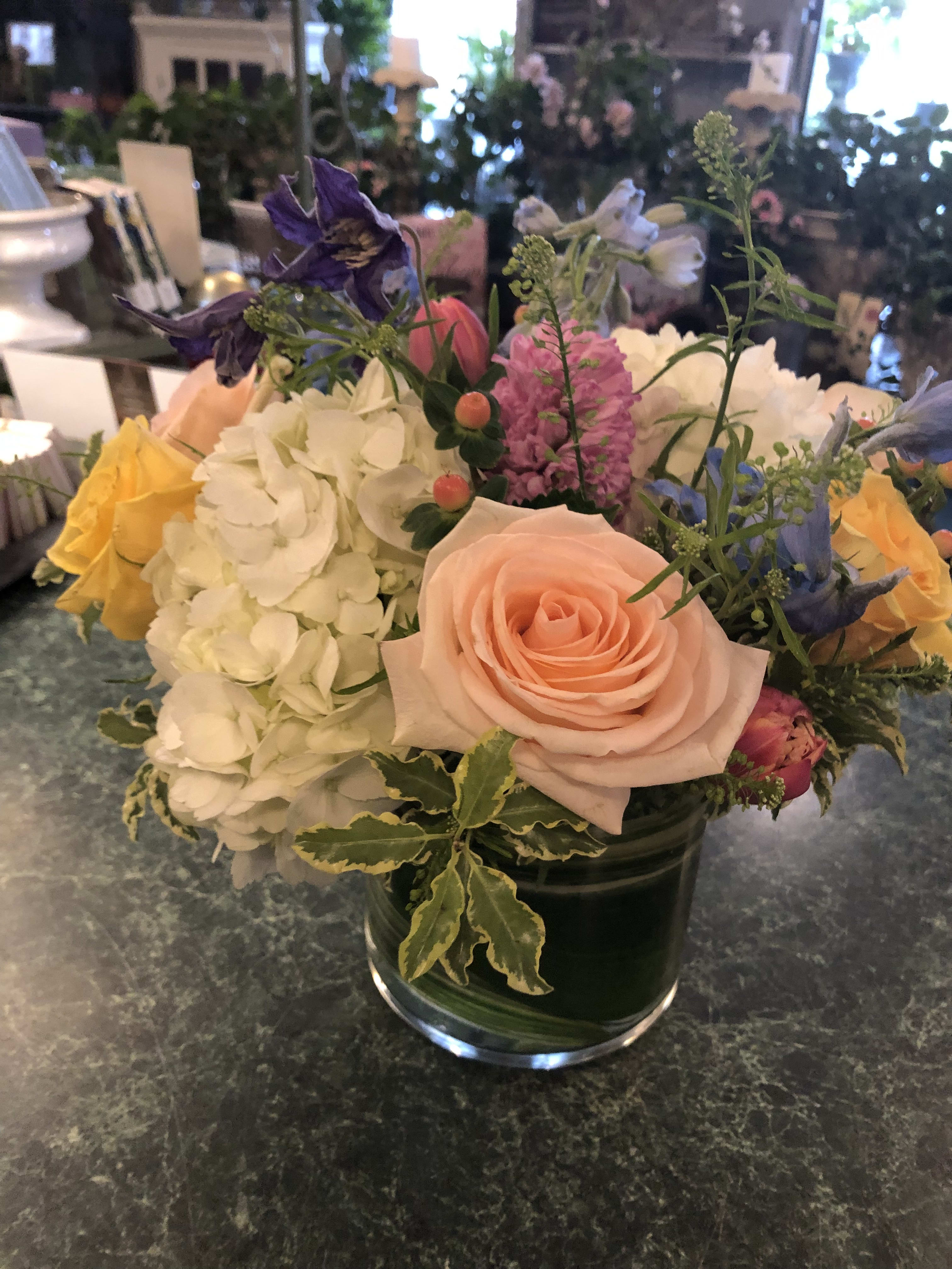 Low glass vase of peach roses, white hydrangeas, and mixed pastel flowers on a green countertop.