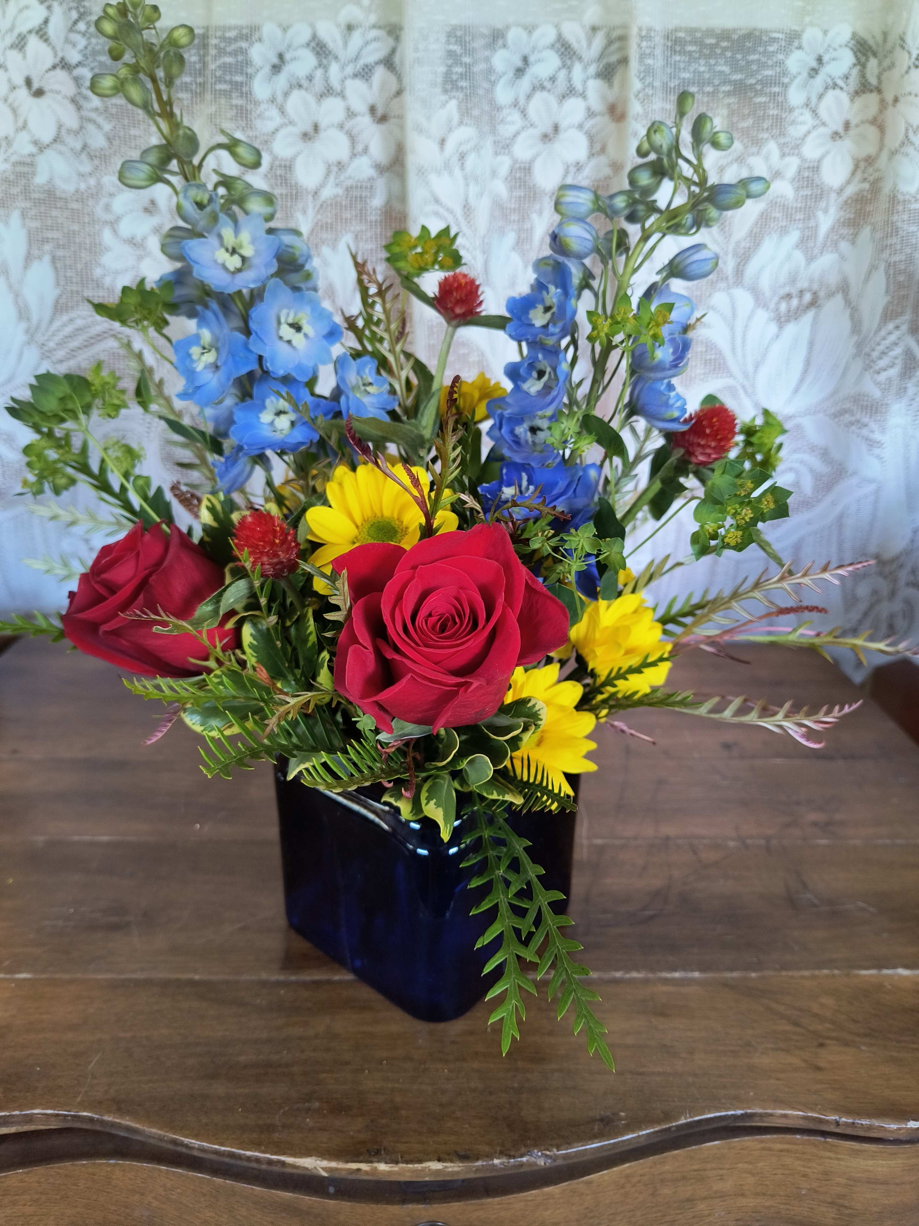 Mixed bouquet of red roses, blue flowers, and yellow daisies in a dark vase