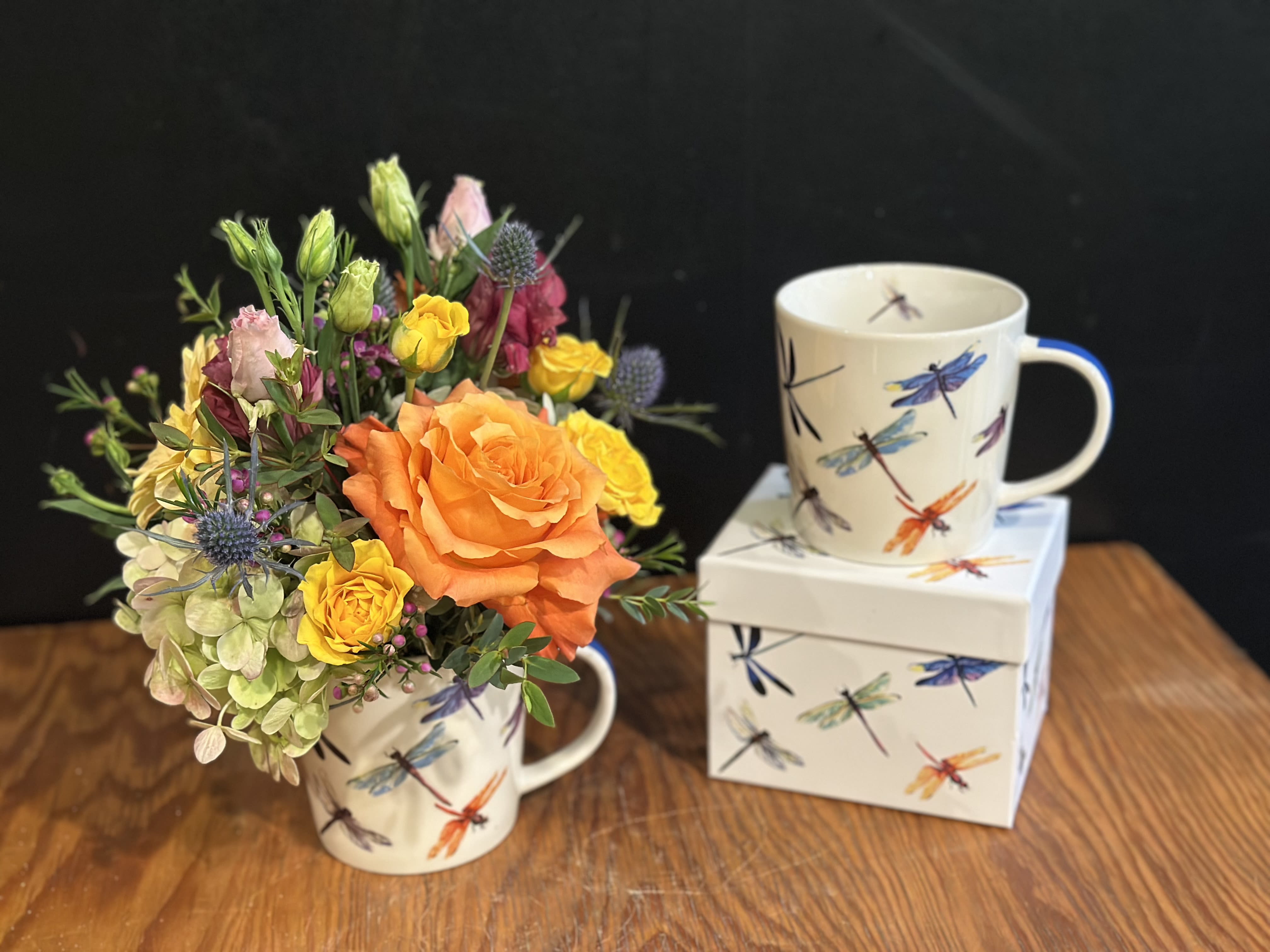 Orange and yellow flower arrangement in a dragonfly mug beside a matching mug and box
