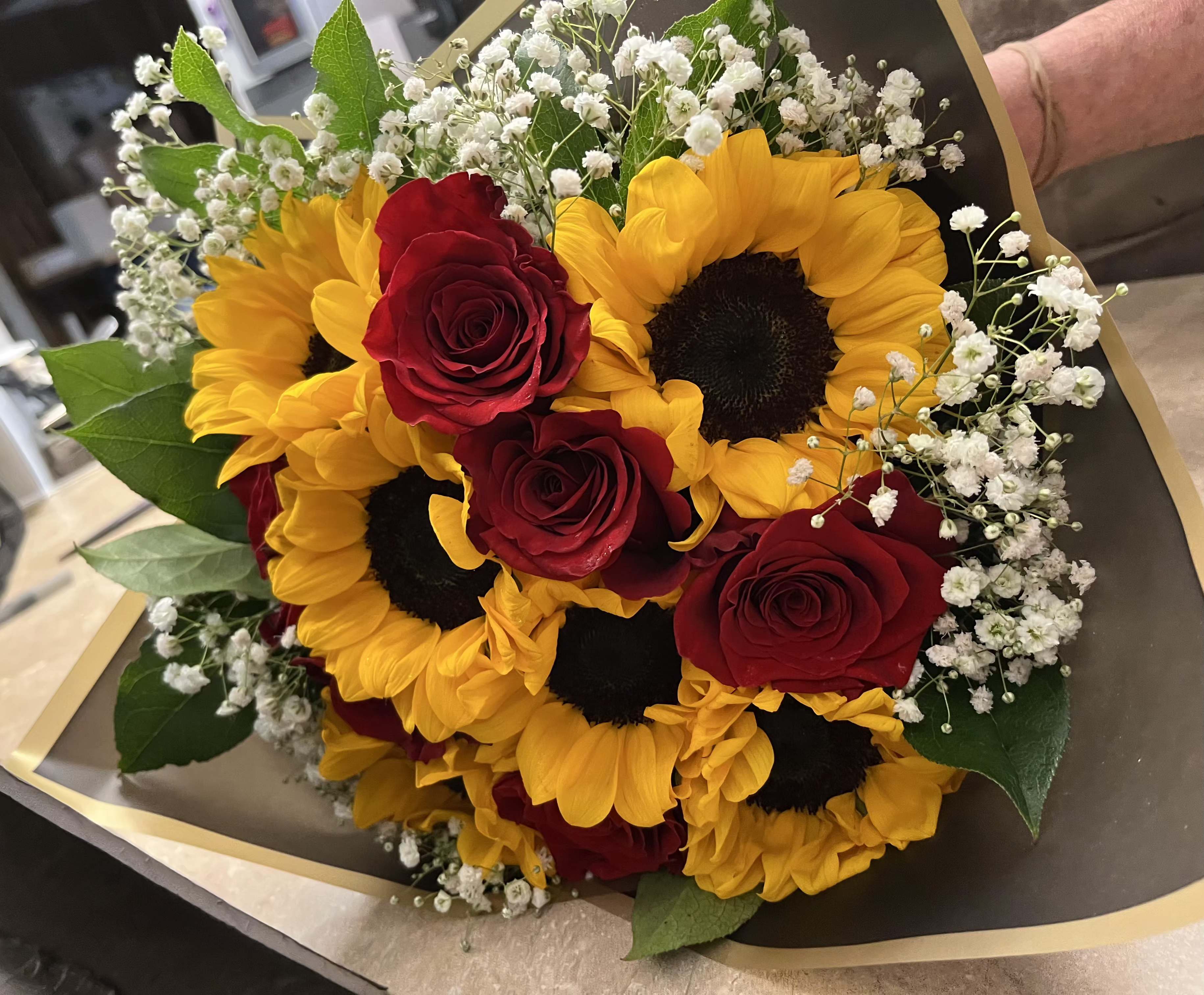 Sunny Love - Bright sunflower faces ring the outside of this hand-tied flower arrangement, making a radiant first impression that feels both cheerful and romantic-perfect as a birthday bouquet, anniversary arrangement, or a warm thank-you gift. The combination of yellow sunflowers, deep red roses, and soft white baby's breath creates a bold, high-contrast look that announces joy while still feeling intimate for the person you're thinking of.  Tightly clustered into a rounded, hand-held shape, the arrangement pairs lush rose centers with airy white accents and fresh green foliage for pleasing texture and depth. Wrapped in black paper with gold trim, the polished, slightly dramatic presentation elevates casual celebrations into memorable moments. Designed to be gifted as shown; no complicated care is required-keep stems in fresh water, trim periodically, and display away from direct heat to extend their vibrant life.