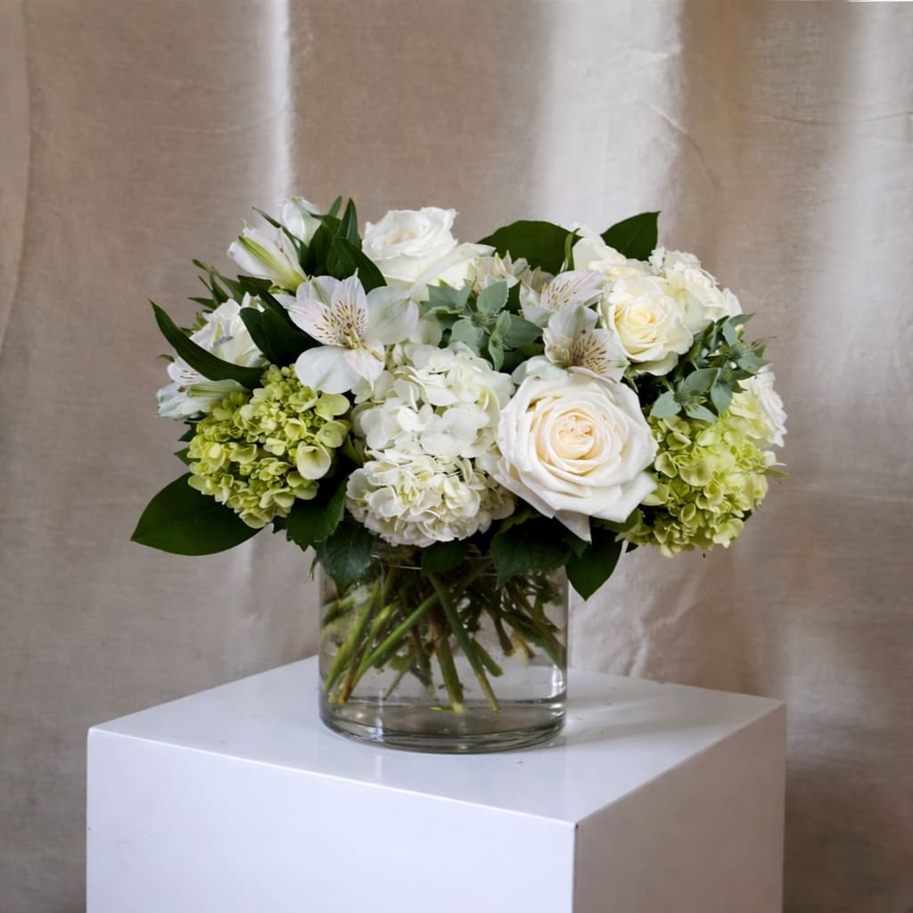 Beach Garden Bouquet - A low, rounded cluster of white roses, creamy white and soft green hydrangeas, and speckled white alstroemeria creates a calm, coastal-inspired flower arrangement perfect as a birthday bouquet, anniversary gift, or a serene centerpiece for Easter or Mother's Day. The Beach Garden bouquet reads as peaceful and refined-the kind of gift that makes the person you're thinking of feel genuinely cared for, whether you're saying "congratulations," "get well soon," or "just because."  Arranged compactly in a clear glass vase, this arrangement sits low on a table or desk without blocking conversation, offering a soft palette of whites and greens that complements spring and summer decor as easily as autumn and winter neutrals. Textural contrast comes from the rounded hydrangea clusters, classic roses, and the delicate, star-shaped alstroemeria-an elegant mix that suits weddings, sympathy, graduations, Father's Day, and holiday gatherings alike. For larger displays, choose the fuller sizes to add extra blooms in the same serene palette; simple care-fresh water and a cool spot-keeps the bouquet looking its best.