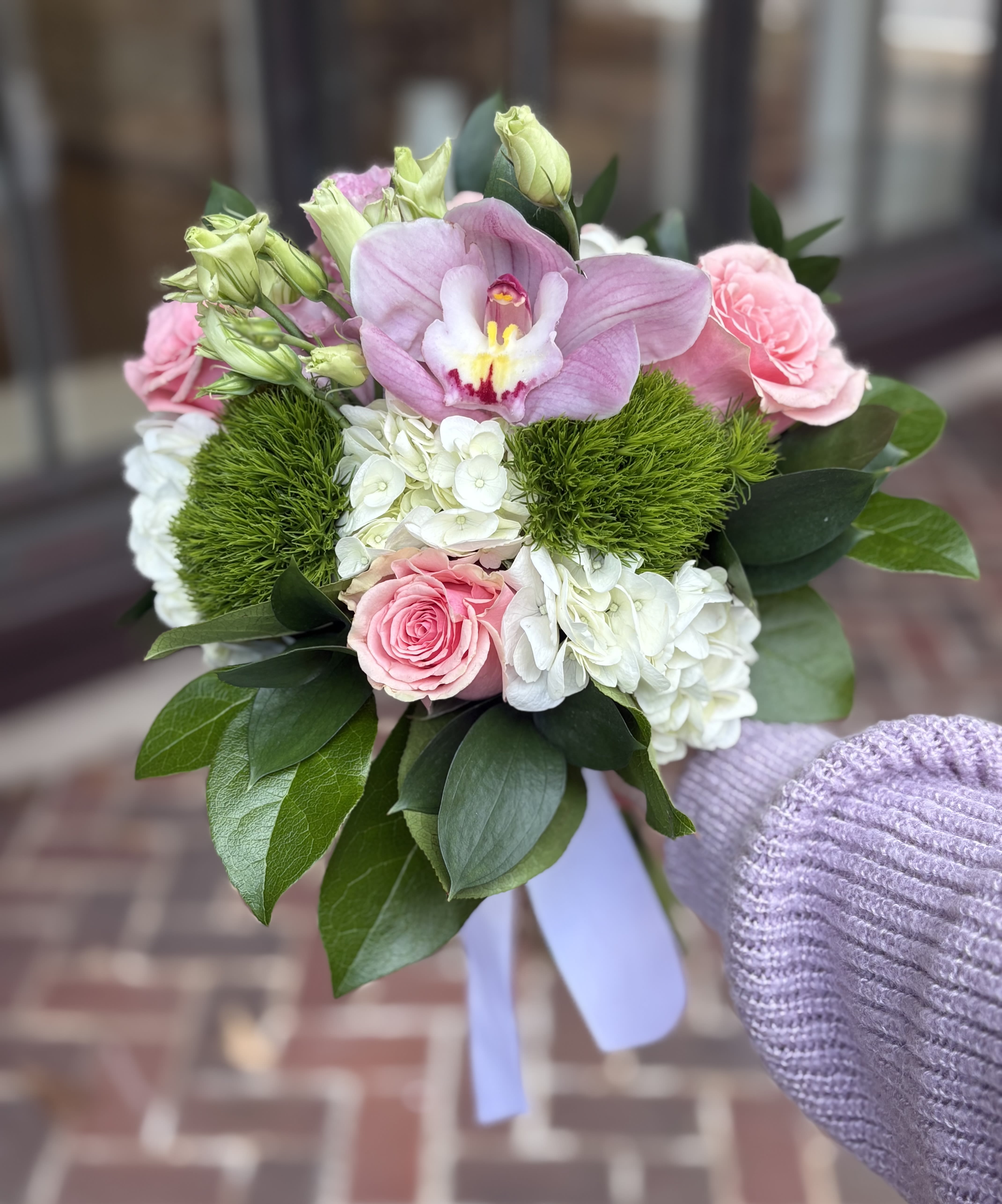 Handheld bouquet of pink orchid, roses, and white hydrangea with green accents and a lavender ribbon.