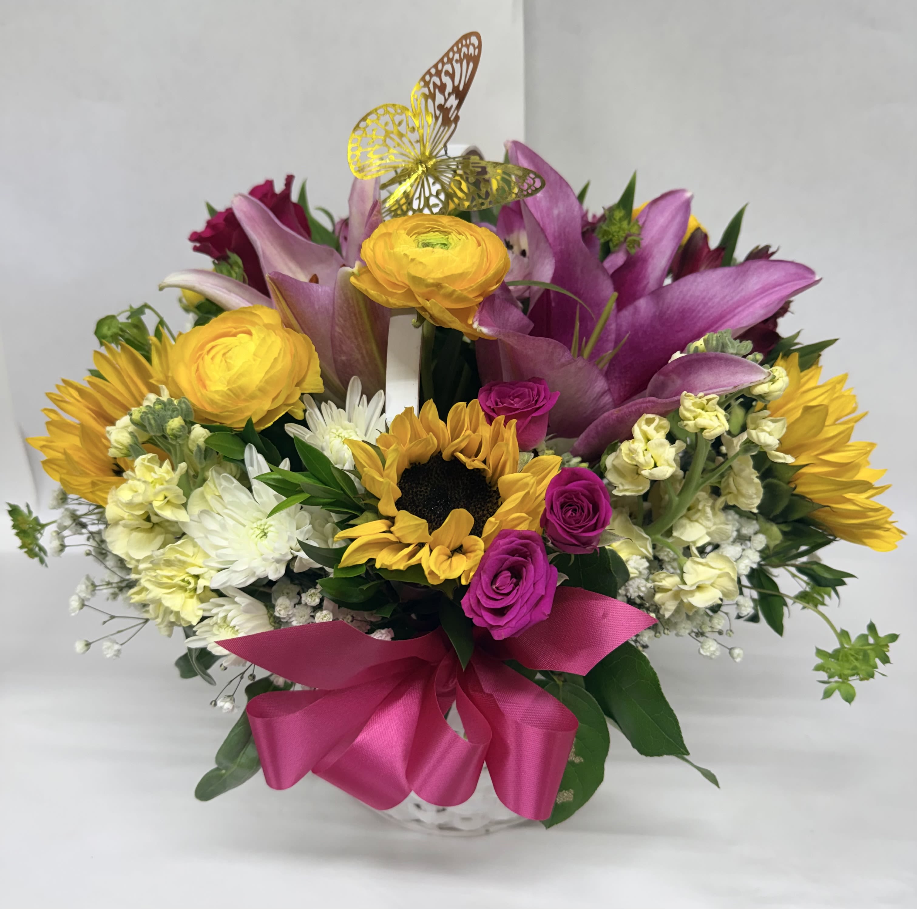 Butterfly Modern Basket - Bright, joyful sunflowers take center stage in this birthday bouquet and celebratory flower arrangement, accented by large pink lilies and yellow roses for a bold, modern contrast. Designed as a cheerful gift for birthdays, congratulations, or just because, the Butterfly Modern Basket uses lively color-warm yellow, cool purple, crisp white-to create an abundant, smile-inducing display that feels festive the moment it's set down.  This rounded composition is arranged in a basket and finished with a vivid pink satin ribbon and a decorative butterfly pick that rises above the blooms for an extra touch of whimsy. White chrysanthemums and fragrant stock add texture and fullness alongside the focal lilies, roses, and sunflowers, giving the arrangement a generous, garden-fresh presence ideal for a mantel, entry table, or office desk.  Perfect for the person you want to make feel celebrated, this arrangement says "you matter" with bold color and playful details-no special occasion required. Sizes are presented at checkout; for best longevity, keep the vase water fresh and out of direct hot sun, and we'll include simple care tips with delivery.