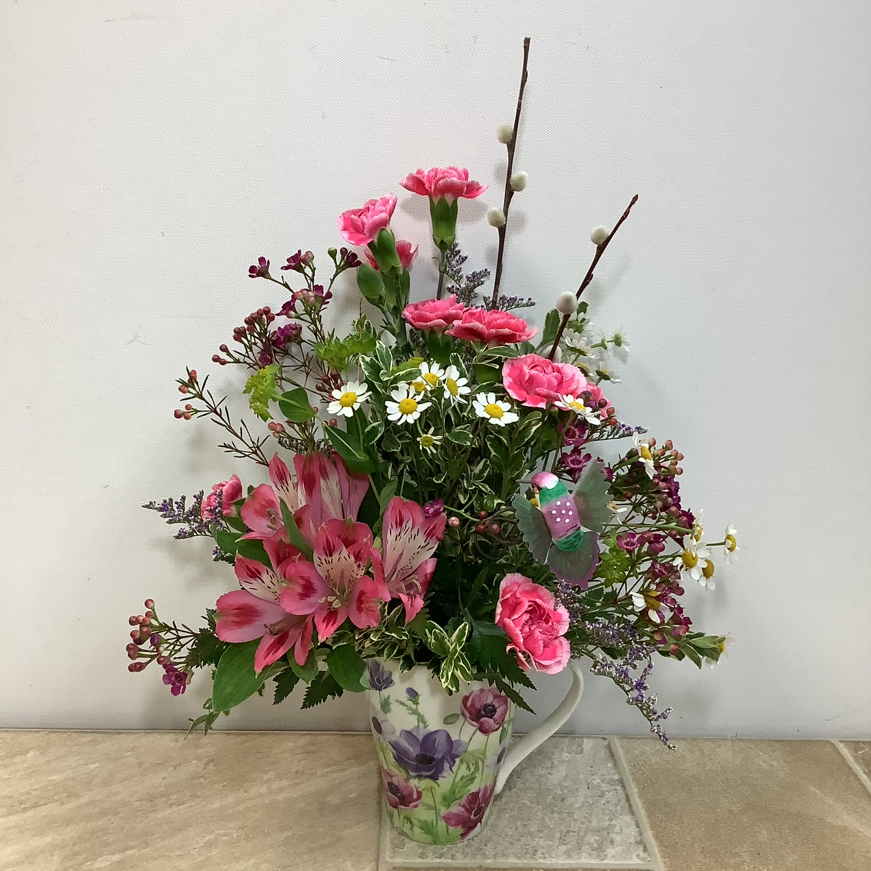 Spring Surprise - Our Spring Surprise bouquet is a cheerful mix of fresh garden blooms beautifully arranged in a pretty ceramic mug. It features feverfew, alstroemerias, mini carnations and wax flower along with pussy willows and a hummingbird. Approximate size: 11” wide x 15” high.  Primary flowers: lily, carnation