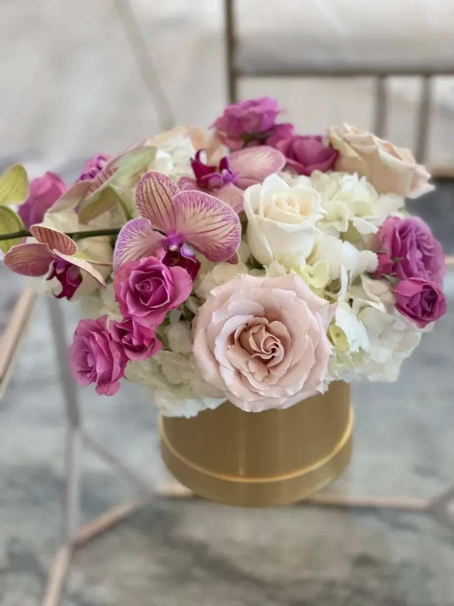 Golden Ticket - A compact cluster of blush, white, purple and peach blooms in a luxe gold round box-this flower arrangement is a romantic gift that reads like a Golden Ticket for anniversaries, birthdays, or "just because" moments. The low, intimate profile and polished color mix make it the kind of floral gift that will make the person you're thinking of feel cherished and seen.  Roses, phalaenopsis orchid sprays, and white hydrangea-like blooms are layered for texture and soft contrast: velvety rose petals and sleek orchid stems sit amid cloud-like hydrangea edges, all contained in a chic gold round box that doubles as decorative keepsake. The compact, horizontal design is ideal for a desk, bedside table, or intimate tabletop display.  Perfect for celebrating a special someone or brightening everyday spaces, this arrangement arrives ready to display-no tall vase needed. Its small, low stature keeps care simple: place in a cool spot out of direct sun and refresh the water source as needed to extend bloom life.