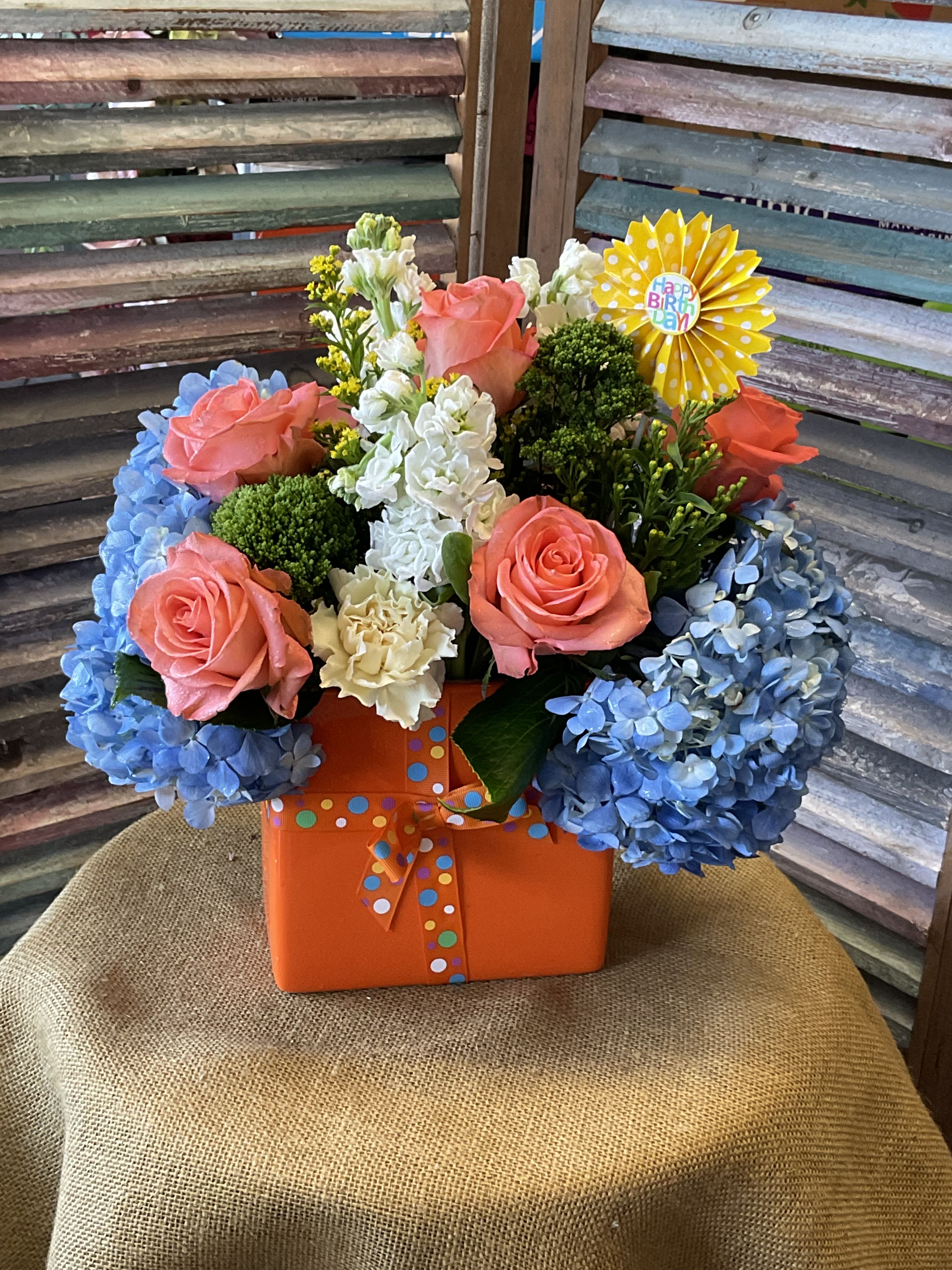 26-121 - Coral-pink roses, blue hydrangeas and white accents arranged in an orange gift box with polka-dot ribbon and birthday pick.