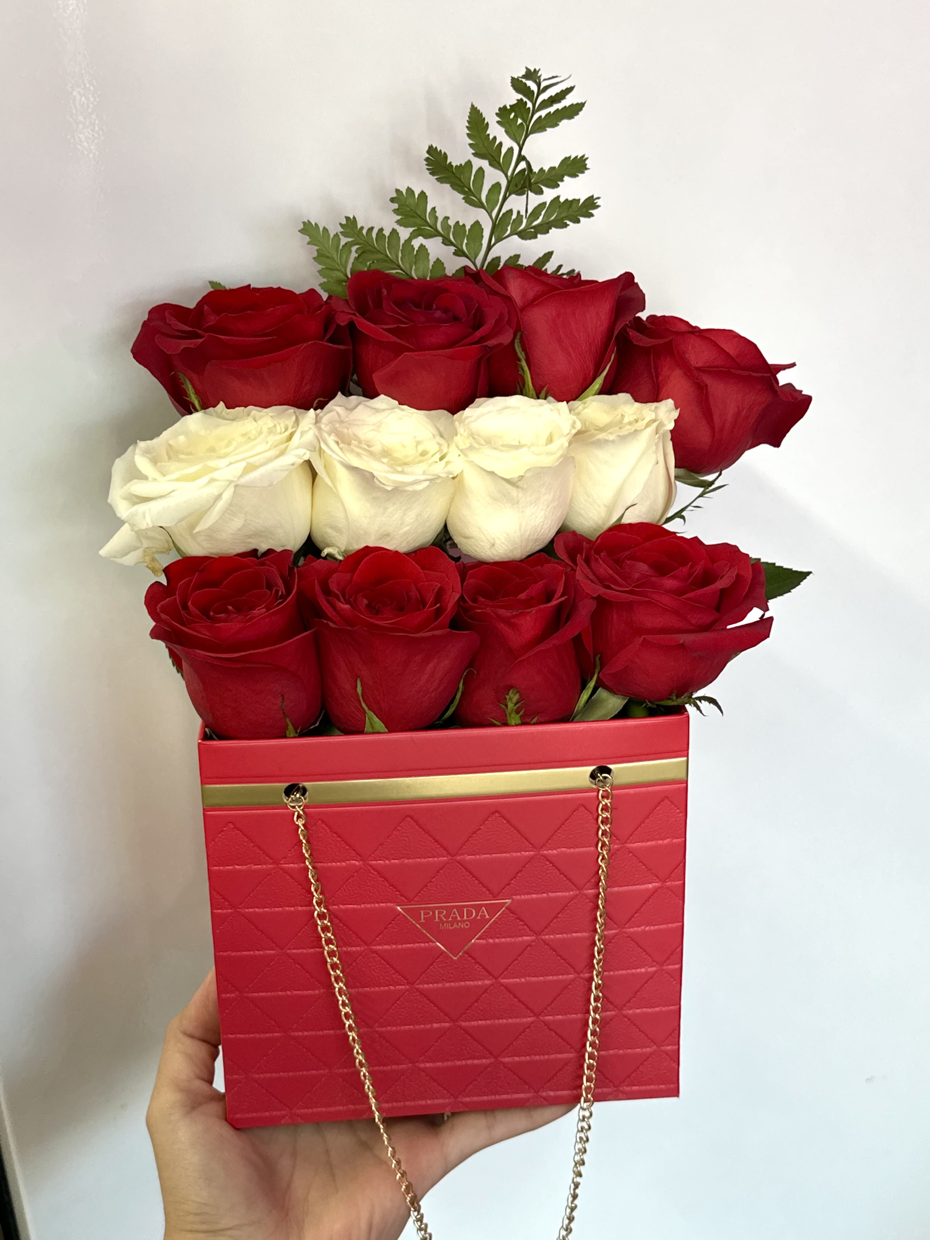 Red and white roses arranged in a pink Prada-style box with a gold chain handle.