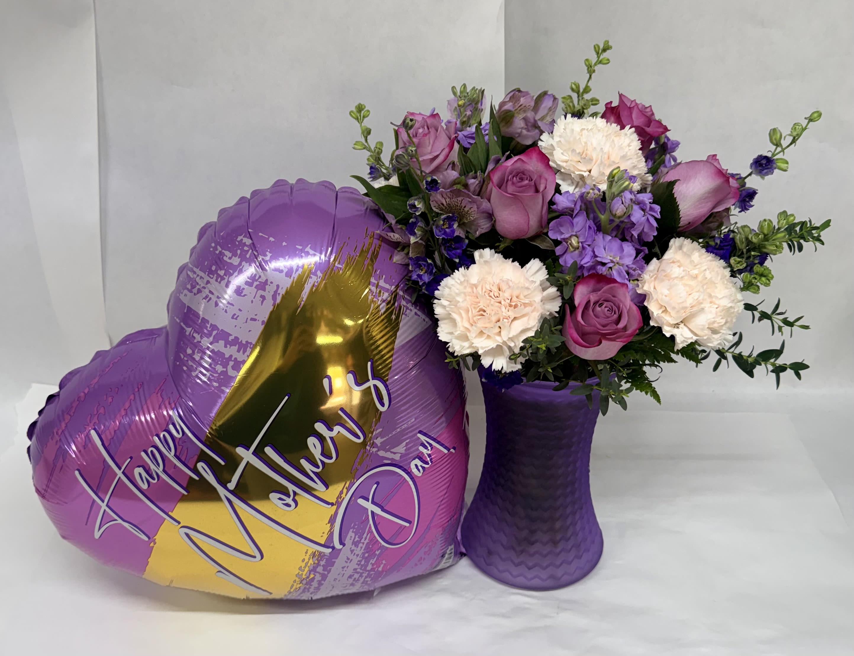 Blooming Beauty Bouquet With Balloon  - Soft lavender and blush roses nestle with pale cream carnations in a textured purple vase, creating a Blooming Beauty bouquet that's perfect as a Mother's Day bouquet or a heartfelt thank-you gift. The large heart-shaped foil balloon reading "Happy Mother's Day" lifts the arrangement into a cheerful, celebratory presentation that will make the person you're thinking of feel seen and appreciated.  This flower arrangement mixes pink and lavender roses, creamy carnations, and small purple accent blooms for a gentle, romantic palette of purple, pink, white, and yellow highlights. Presented in a sturdy purple vase, the bouquet balances lush petals and delicate texture for a display that looks full without being fussy. The included heart balloon adds height and a festive note, making the gift ready to present straight from delivery.  Made to honor a mother, grandmother, or anyone who deserves special recognition, this arrangement suits a brunch table centerpiece or a bedside surprise that brightens the whole day. Order with confidence for same-week delivery where available; to keep blooms looking their best, place in fresh water away from direct sun and drafts and refresh water every two days.