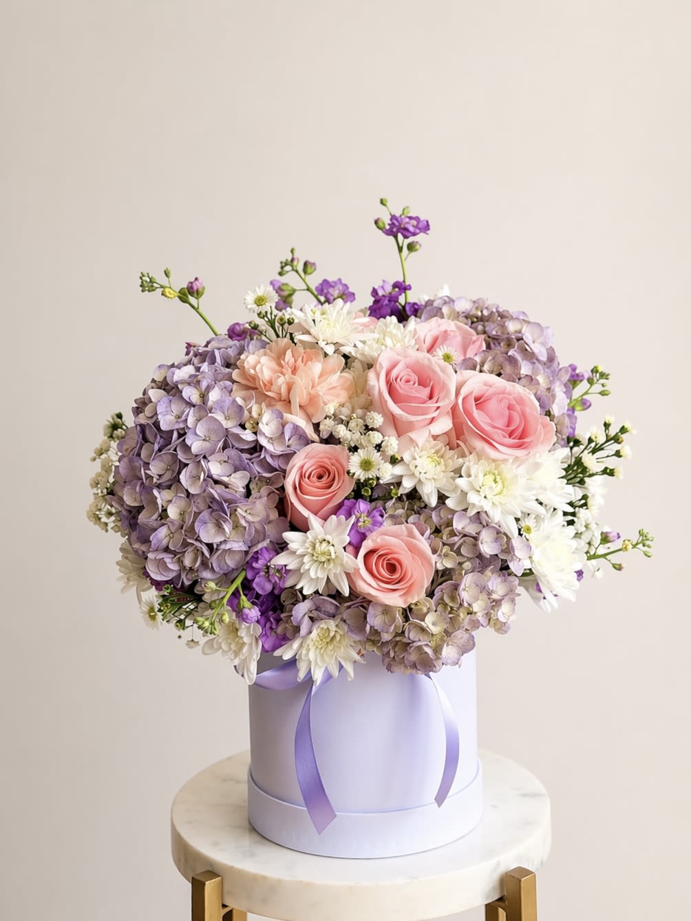 Blush Lavender Box - A lush cluster of pink roses and lavender hydrangeas makes a soft, romantic statement in this blush lavender box-ideal as a birthday bouquet, anniversary arrangement, or Mother's Day gift. The pastel mix of pink, purple, white and peach reads feminine and celebratory, the kind of floral gift that will make the person you're thinking of feel genuinely cherished and seen.  Roses, full lavender hydrangeas, white daisy-like blooms and gentle peach accents are packed tightly into a round, pale lavender hat box and finished with a matching lavender ribbon for a polished, gift-ready presentation. The design's dense, rounded silhouette gives abundant visual impact from every angle, with varied textures from the hydrangea's cloud-like heads to the crisp centers of the daisies.  Perfect for sending warm congratulations, heartfelt thanks, or a thoughtful just-because surprise, this arrangement arrives ready to display-no vase required. Care is simple: keep the box in a cool spot away from direct sun and replenish water as needed to maintain freshness for several days.
