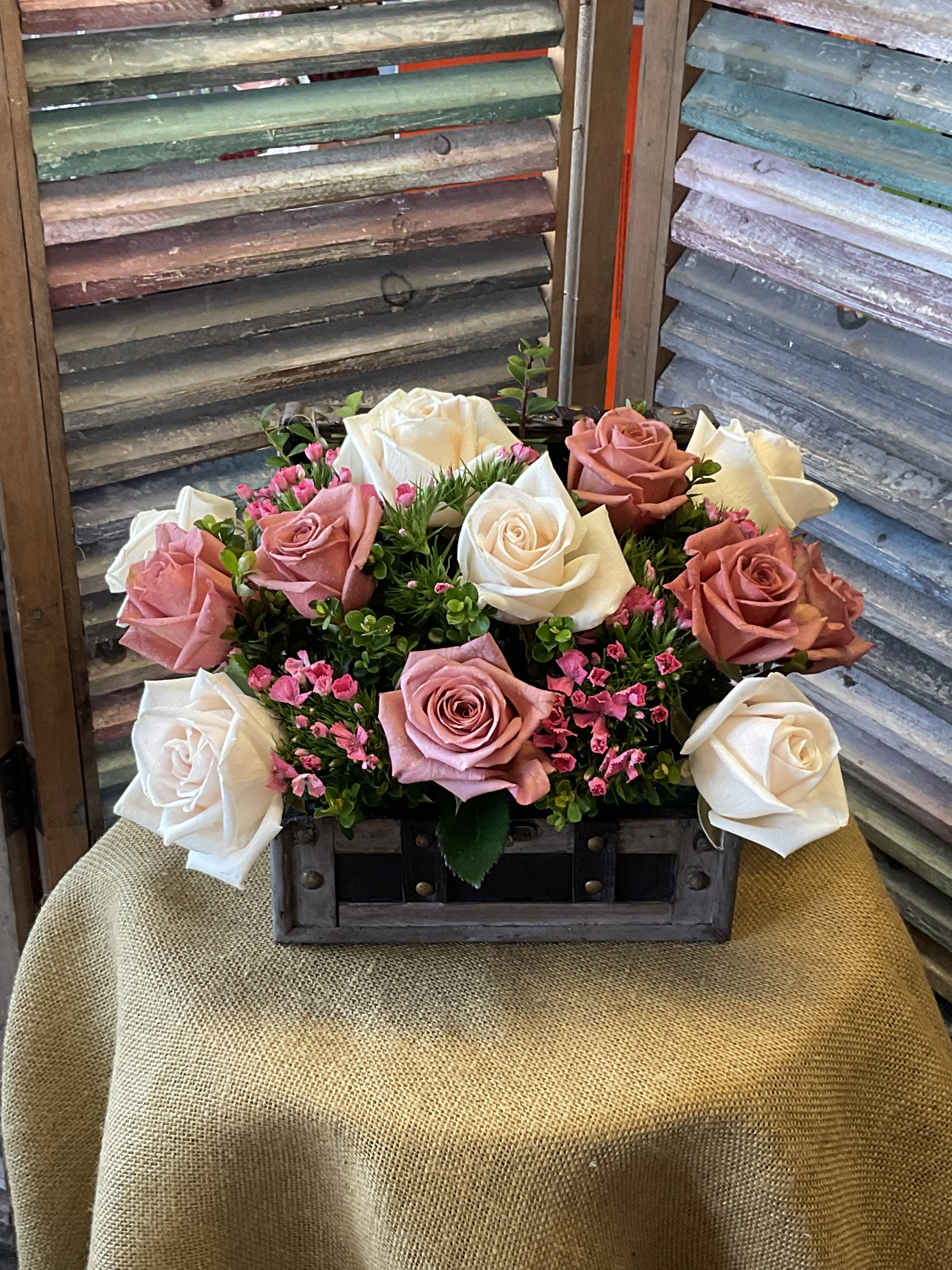 26-120 - Cluster of pink and white roses with petite pink fillers and greenery, arranged in a rustic wooden crate.
