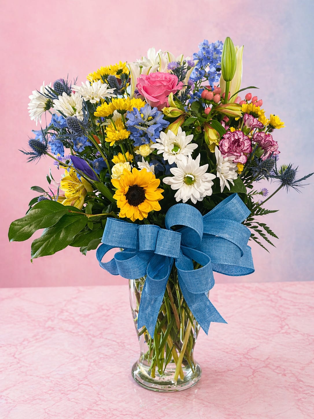 Blue Skies and Sunshine - Bright, cheerful, and full of life—Blue Skies &amp; Sunshine is a feel-good arrangement that brings instant happiness. Featuring sunny sunflowers, crisp white daisies, soft pink roses, and pops of blue delphinium, this bouquet is tied together with a beautiful blue bow for that extra touch of charm. Designed in a clear glass vase with lush greenery, it’s perfect for celebrations, get well wishes, or simply sending a little joy “just because.” Each arrangement is uniquely crafted with fresh, seasonal blooms—because every moment deserves something special, delivered with love.