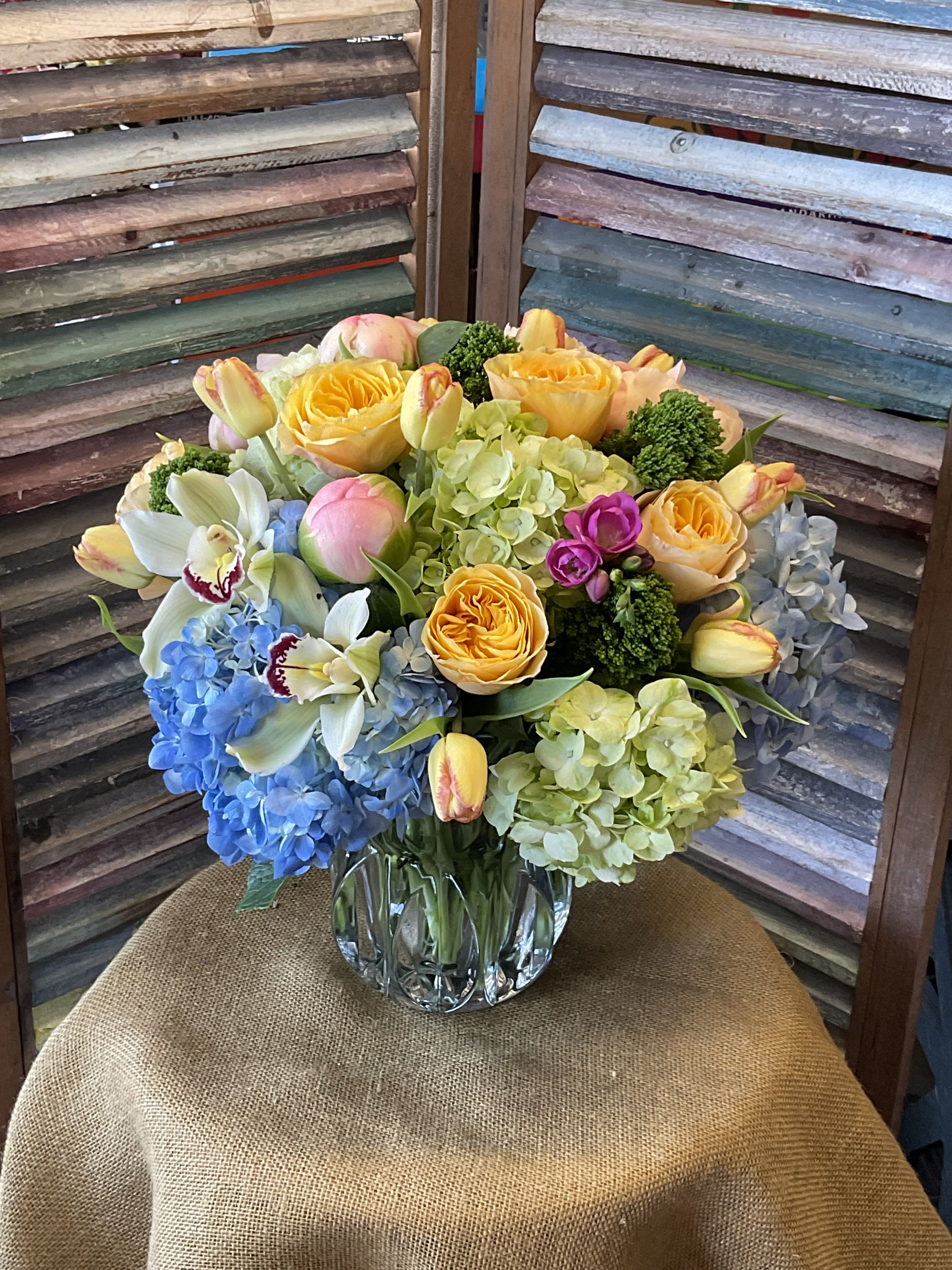 26-123 - Lush rounded bouquet of yellow garden roses, blue &amp; green hydrangeas, peonies, orchids, freesia and tulips in a beautifully designed clear vase that can be reused as a decorative keepsake. 
