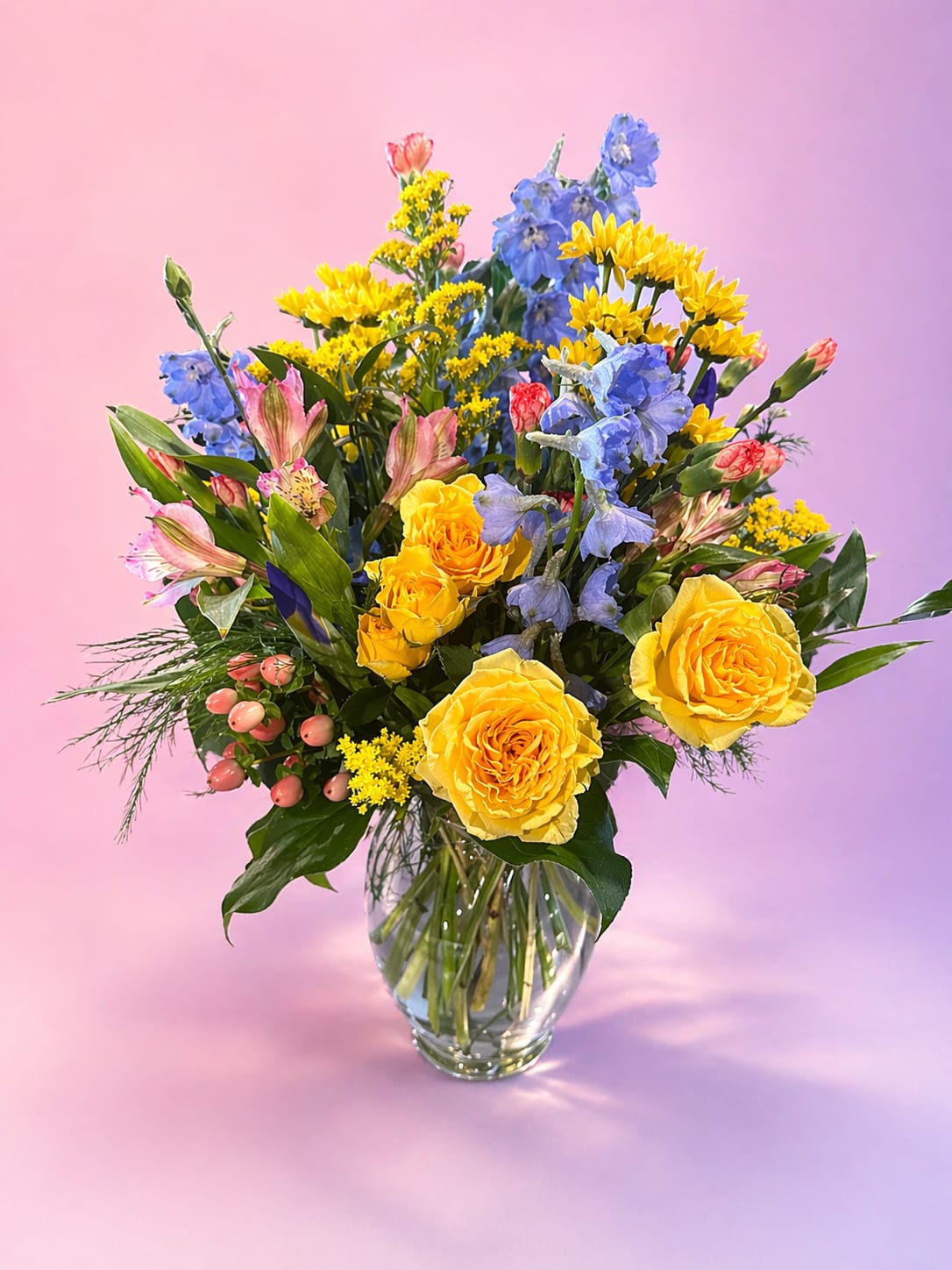 Golden Hour Garden - Warm, radiant, and effortlessly beautiful—Golden Hour Garden captures the glow of a perfect sunset in bloom. Featuring lush golden roses, soft blue delphinium, cheerful yellow daisies, and delicate pink accents, this arrangement is full of movement and natural charm. Designed in a clear glass vase with airy greenery and seasonal textures, it feels fresh-picked and full of life.