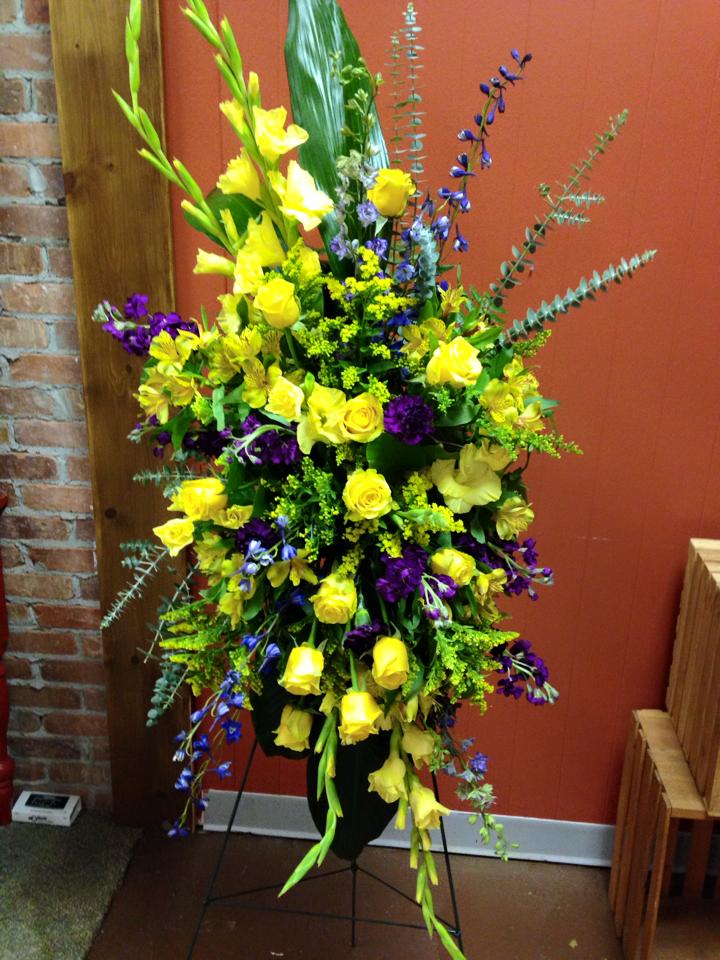 Tall floral arrangement with yellow roses and purple flowers in a black stand
