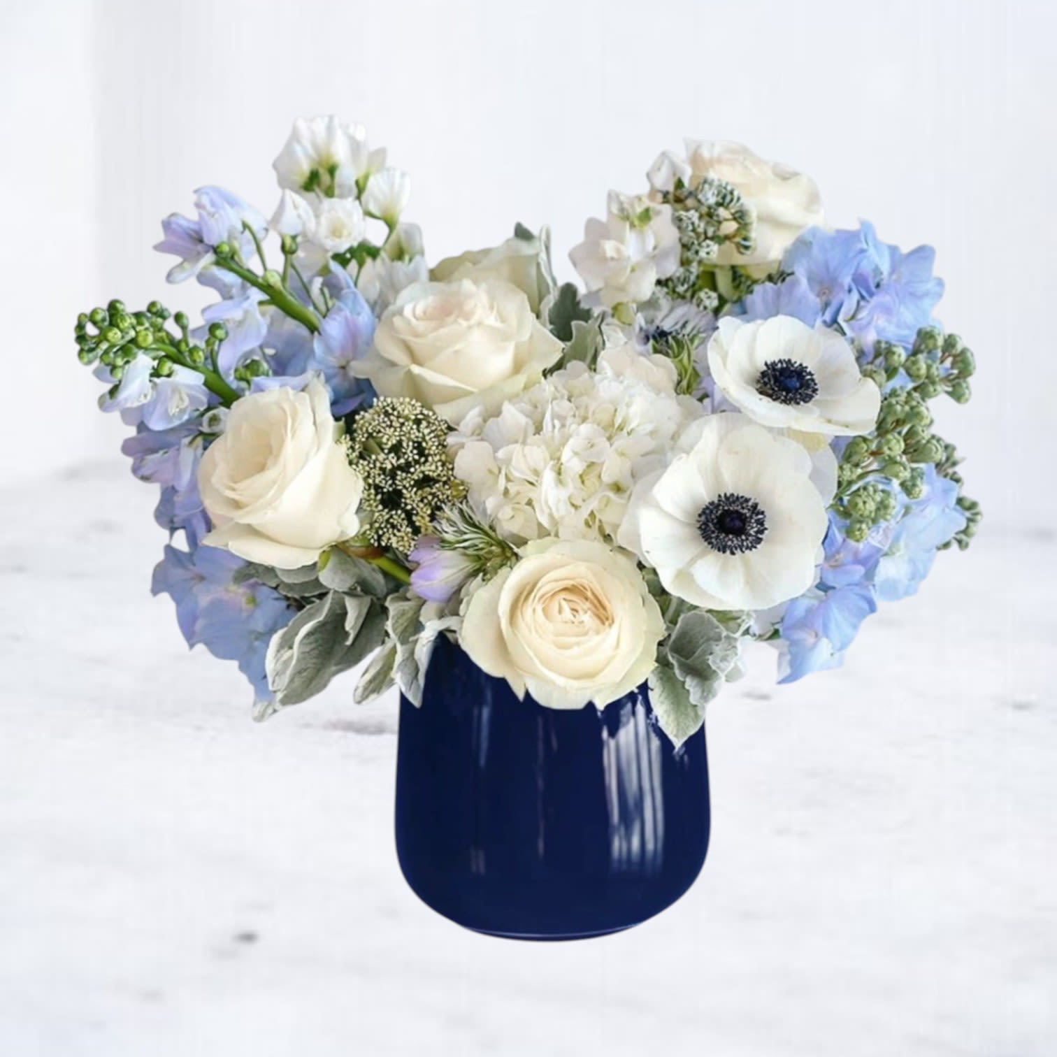 Ink &amp; Ivory - A quiet bouquet of white and soft-blue blooms designed to comfort the person you're thinking of: this flower arrangement for "I'm Sorry" moments mixes white roses and anemones with pale blue delphinium and clusters of white hydrangea for a soothing, refined look. The contrast of the airy, pale flowers against a glossy dark blue ceramic vase gives the arrangement a modern, calming presence-perfect for offering sympathy, sending a gentle "thinking of you" note, or welcoming a new baby with understated elegance.  Creamy roses and white anemones with their dark centers provide focal clarity, while the soft delphinium adds vertical movement and the hydrangea brings full, textural volume. The rounded dark-blue vessel anchors the palette and reads contemporary on a mantel, bedside table, or nursery dresser. Size and proportions are crafted to feel intimate yet substantial enough to convey care without overwhelming a small space.  Send this arrangement as a thoughtful way to say you care-whether to comfort someone after a loss, to let a friend know they're in your thoughts, or to celebrate the arrival of a newborn with serene beauty. Care: keep blooms in fresh water, trim stems slightly and remove any foliage below the waterline; replace water every other day to extend freshness. Delivery available in local service areas.