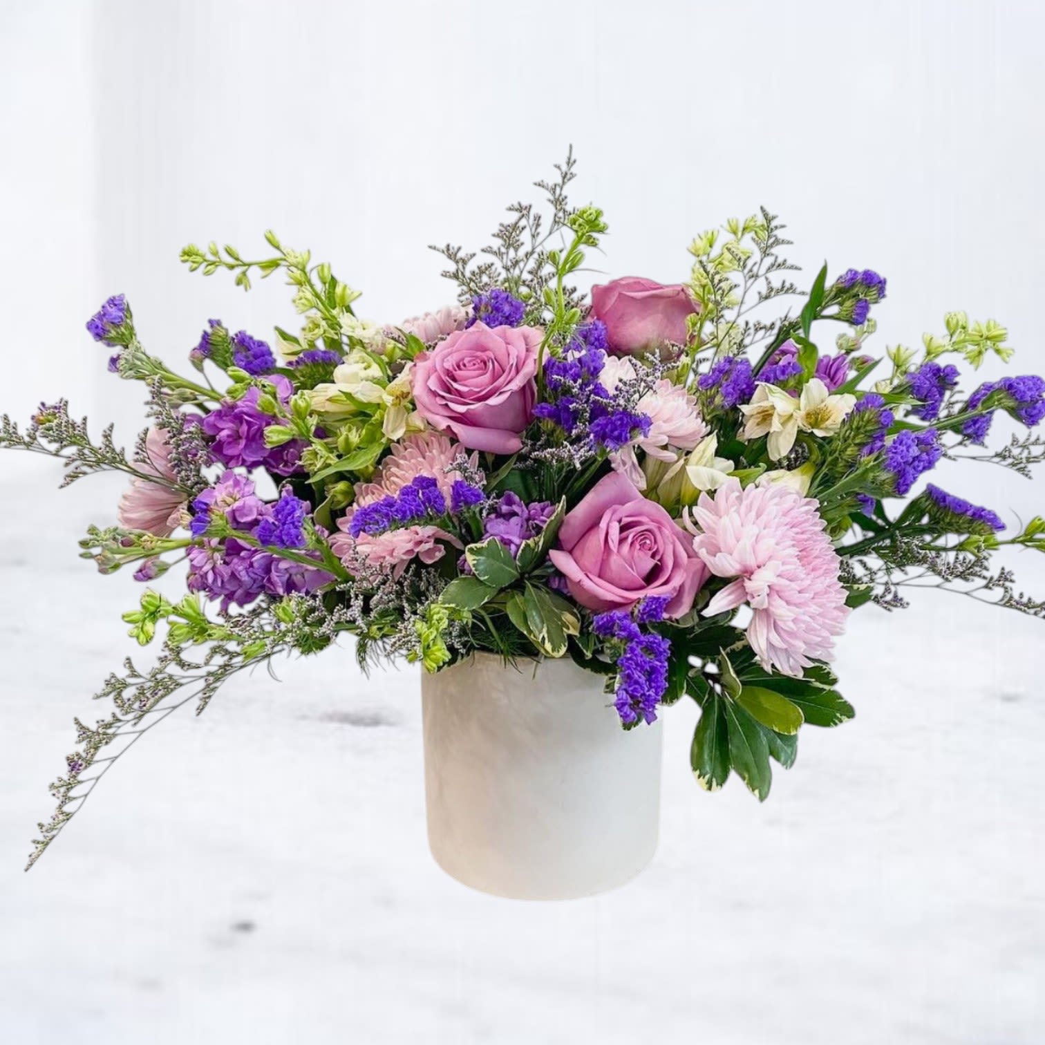 Lavender roses and purple flowers in a white cylindrical vase