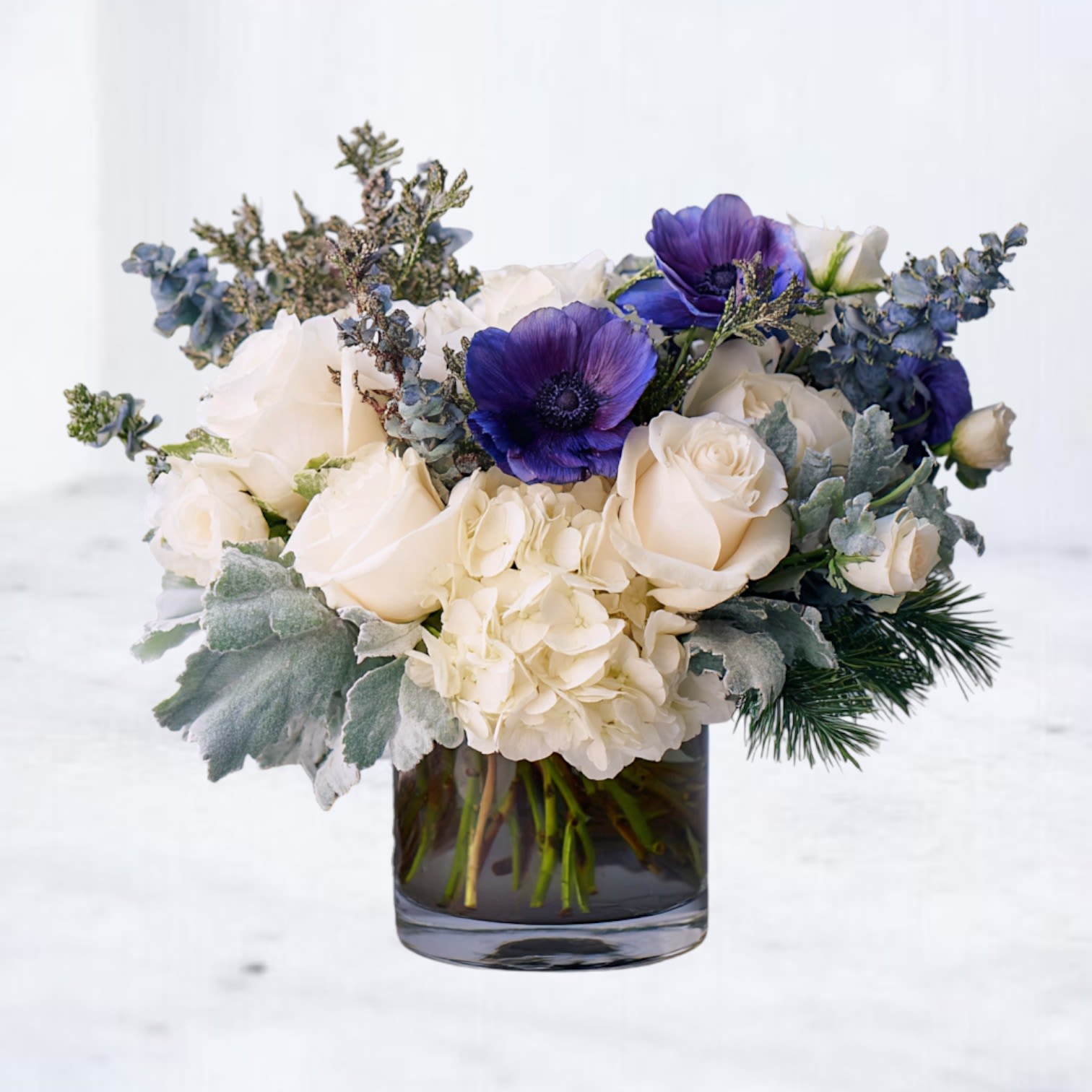 Low arrangement of white roses, hydrangeas, and purple anemones in a clear glass cylinder vase