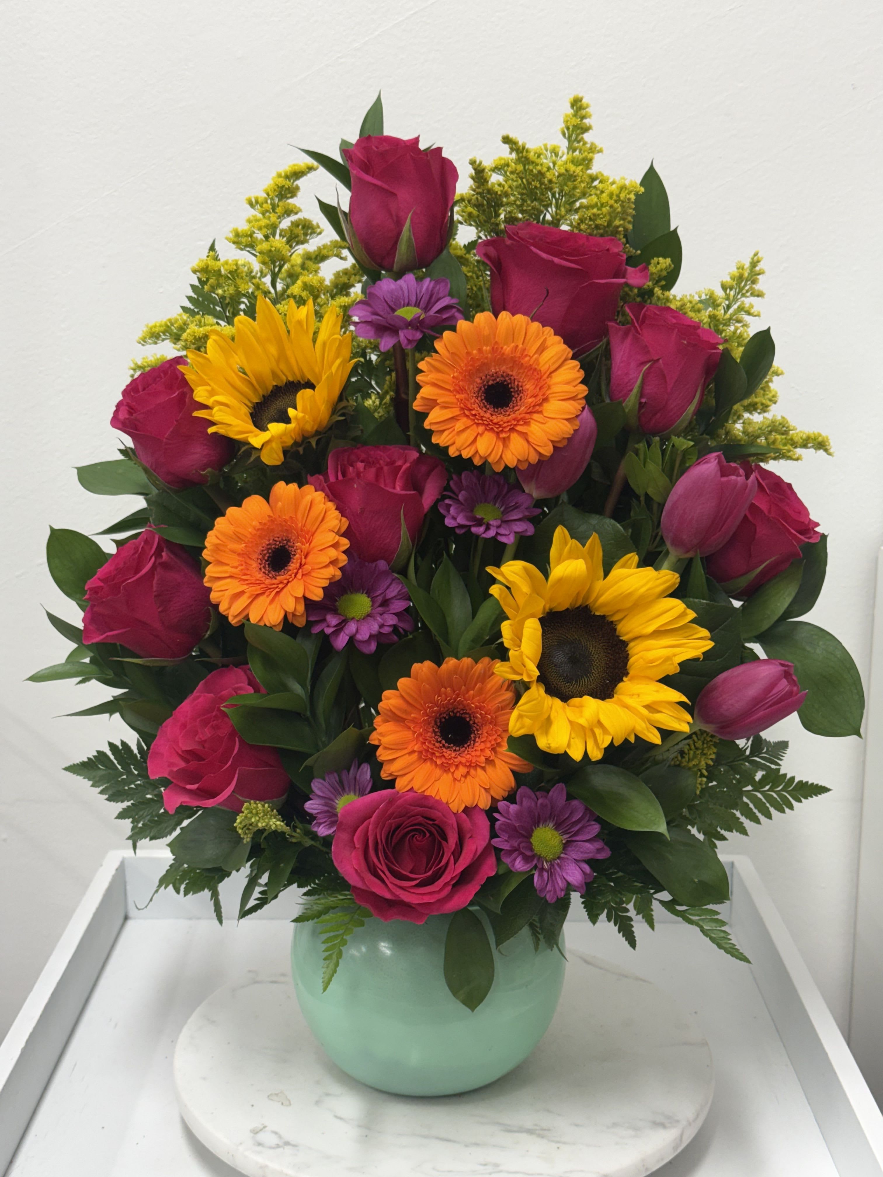 Aromatic Harmony - Rounded bouquet of hot pink roses, yellow sunflowers, orange gerberas and purple daisies in a mint ceramic vase.