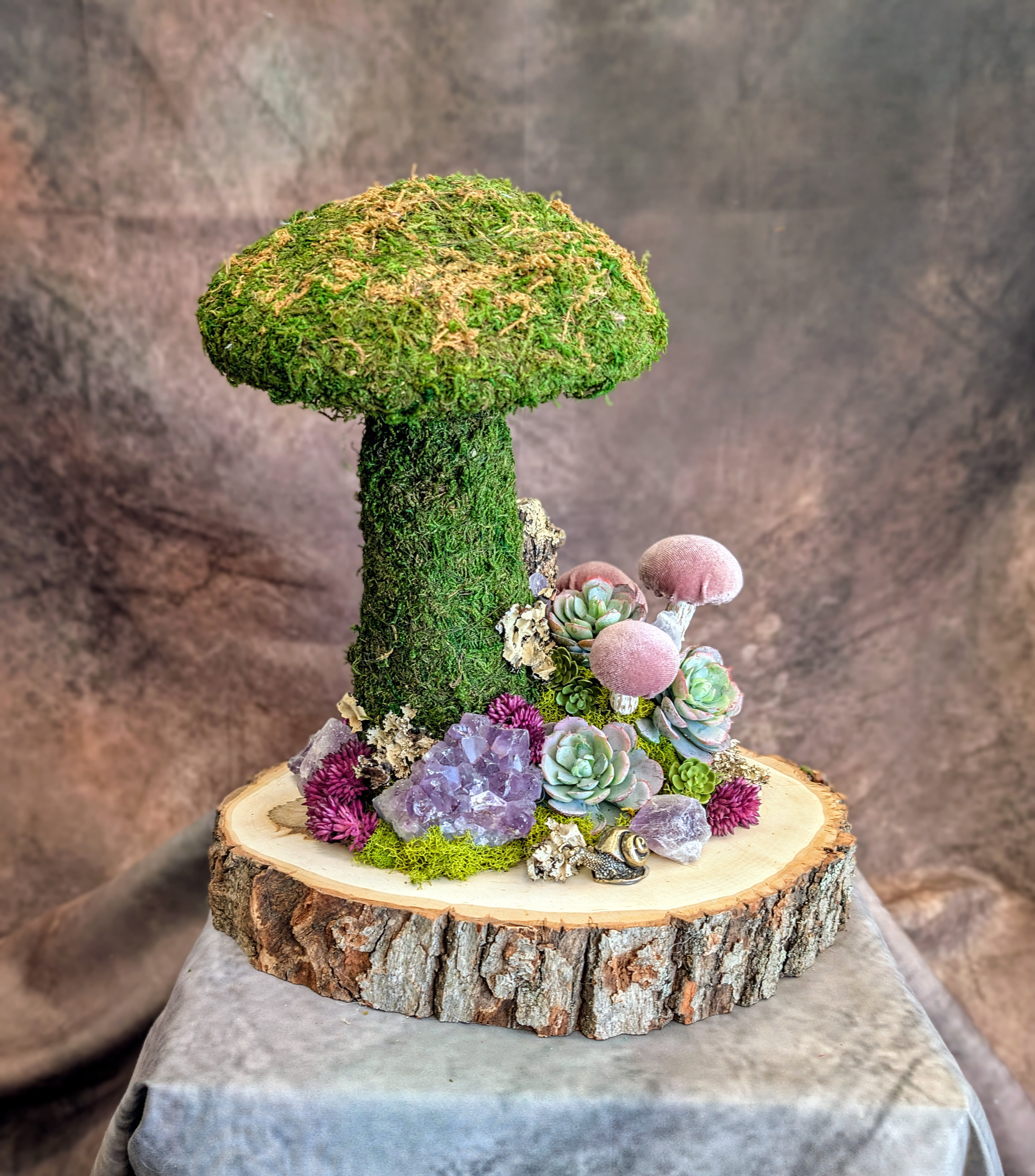 Bramble- Preserved And Permanent Botanical Arrangement - An enchanting woodland vignette, Bramble brings the quiet magic of the forest floor into a refined, sculptural design. A lush, moss-covered mushroom rises from a natural wood slice base, surrounded by a curated garden of succulents, textural botanicals, and shimmering amethyst crystal clusters.  Soft sage and dusty blue echeveria mingle with rich plum accents and preserved moss, creating a layered palette that feels both organic and elevated. Delicate details—miniature mushrooms, natural lichen, and stone—add depth and a sense of story, like a hidden corner of an enchanted grove.  Handcrafted at Windmill Floral Expressions in Las Vegas, each piece is thoughtfully composed and completely one-of-a-kind. Designed as a long-lasting botanical art piece, Bramble is perfect for gifting or adding a touch of mystical, nature-inspired décor to your space.  Available for same-day delivery in Las Vegas and Henderson, this design is created fresh to order using premium materials and our signature artistic style.  This arrangement measure sapproximately 13 inches high by 12 inches wide.