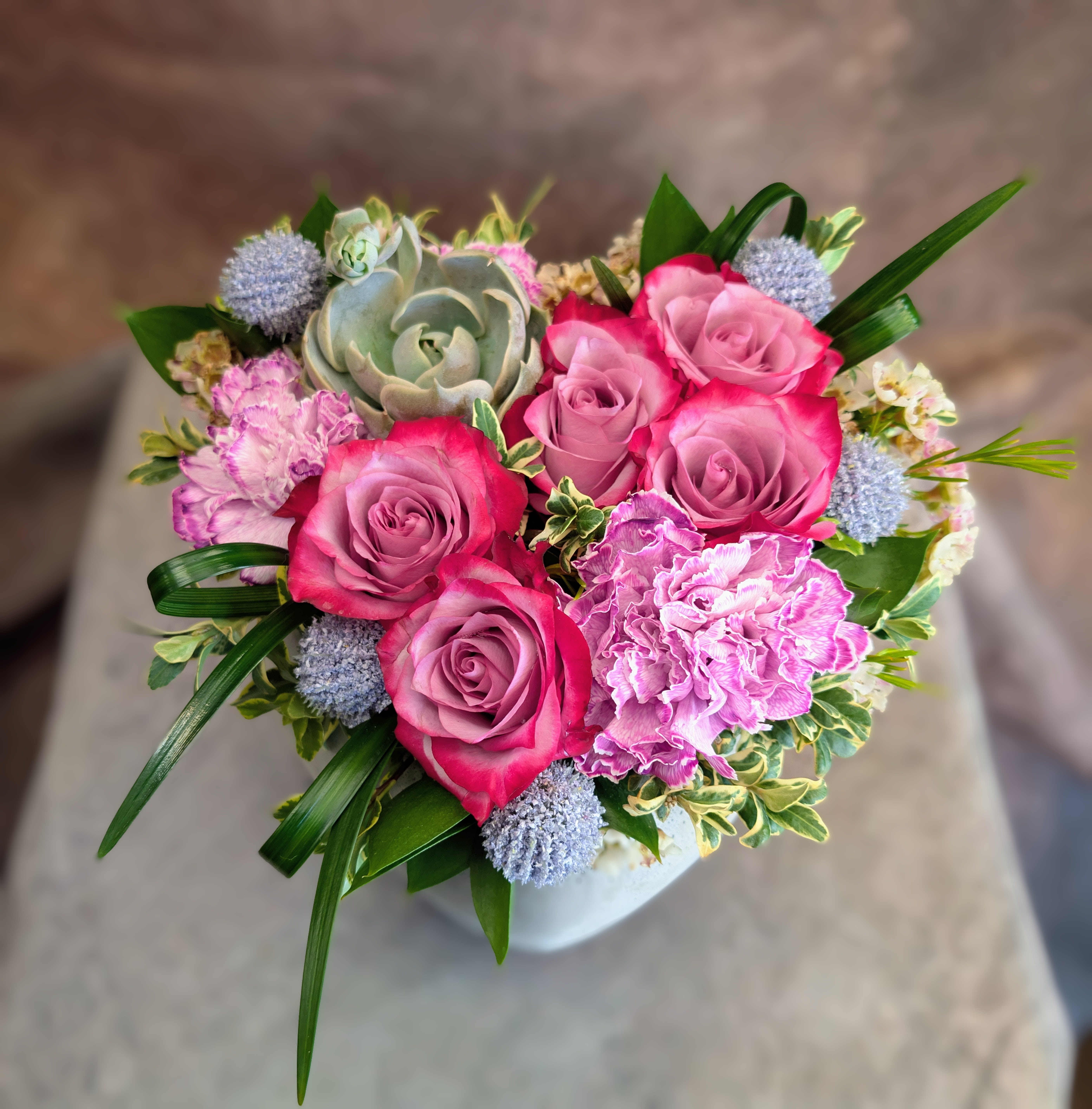 Posy - Softly romantic and thoughtfully designed, Posy is a modern heart-shaped flower arrangement in Las Vegas that blends garden charm with contemporary style. Arranged in a matte heart-shaped concrete container, this petite design offers a meaningful, elevated take on a classic floral gesture.  Bi-color roses in luminous lavender and fuchsia tones create the focal point, their layered petals adding depth and richness. They are complemented by lavender and white variegated carnations, bringing soft texture and movement to the composition. Cool-toned accents and fresh greenery provide balance, while a sculptural echeveria succulent adds a subtle, modern edge.  Measuring approximately 8.5 inches at its widest, Posy is designed in a compact, full style—perfect for intimate gifting without sacrificing impact. The contrast of soft florals against the clean, natural texture of the concrete vessel gives this piece a refined yet approachable feel.  Handcrafted at Windmill Floral Expressions in Las Vegas, each arrangement is made to order using premium, fresh blooms. We do not offer pre-made designs, ensuring every piece is one-of-a-kind and created with care.  Available for same-day flower delivery in Las Vegas and Henderson, Posy is a beautiful way to send something personal, romantic, and thoughtfully designed.