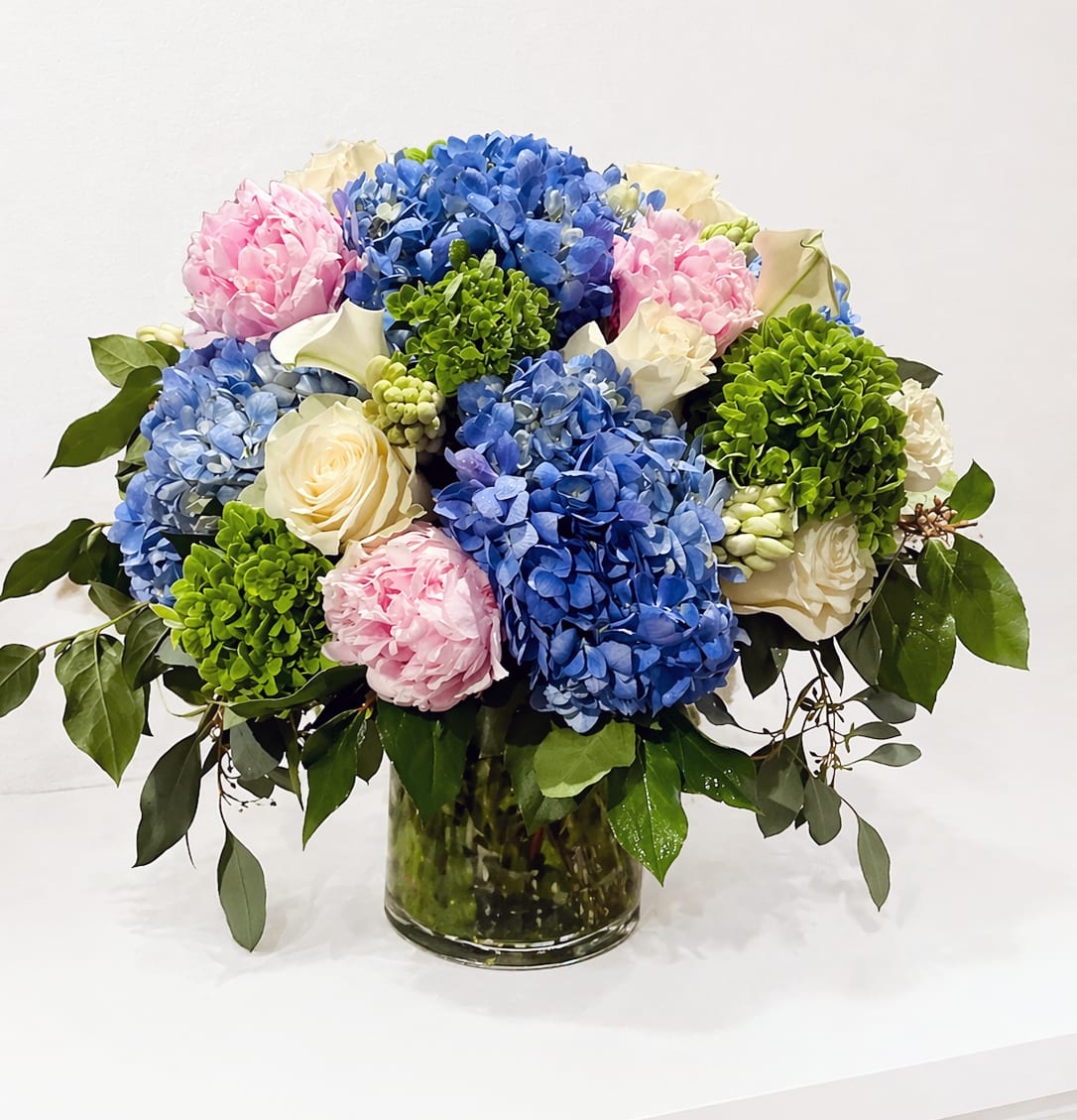 Mom's Garden Elegance - Celebrate her with something as graceful and nurturing as she is. Mom's Garden Elegance is a soft, refined arrangement designed to honor love, care, and timeless beauty. Lush blue hydrangea create a cloud-like foundation, layered with delicate blush peonies, creamy white roses, elegant calla lilies, and fresh green accents for a naturally abundant feel.  Arranged in a clear glass cylinder, this piece blends classic elegance with a modern, airy touch—perfect for brightening her home, her office, or the center of a special Mother’s Day table.  Style: Soft, elegant, garden-inspired Color Palette: Blue, blush pink, ivory, and fresh green Vase: Clear glass cylinder Perfect For: Mother’s Day brunch, home décor, or a heartfelt surprise delivery  A beautiful way to say thank you, Mom—for everything she’s done and everything she is.