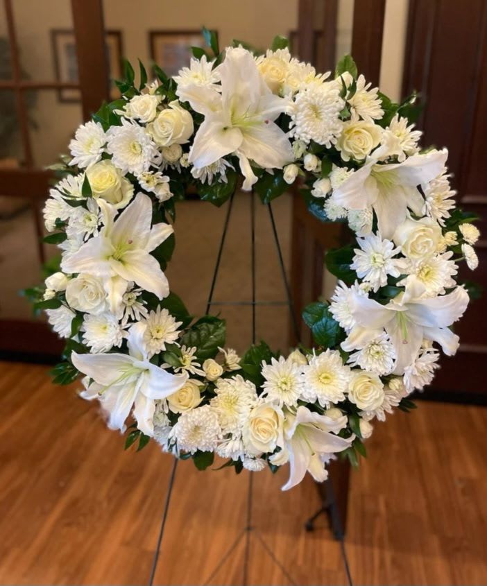 Heavenly Grace Round Wreath - A serene circular sympathy wreath designed to offer calm and grace, this funeral flower arrangement immediately reads as a respectful tribute for a memorial or celebration of life. The round shape and pale palette of white lilies and cream roses create a gentle focal point, while clusters of white daisies and chrysanthemums add soft texture-an arrangement that lets the person you're honoring feel seen and remembered.  Densely packed blooms form a full, layered ring set against deep green foliage for crisp contrast and balanced structure. Large white lilies, ivory roses, and daisy-like chrysanthemum clusters are arranged to create a refined, formal silhouette; the wreath arrives ready on a metal stand for easy display. Its understated white, green, and pale cream tones suit funeral services, graveside memorials, or any solemn occasion calling for classic elegance and quiet comfort.