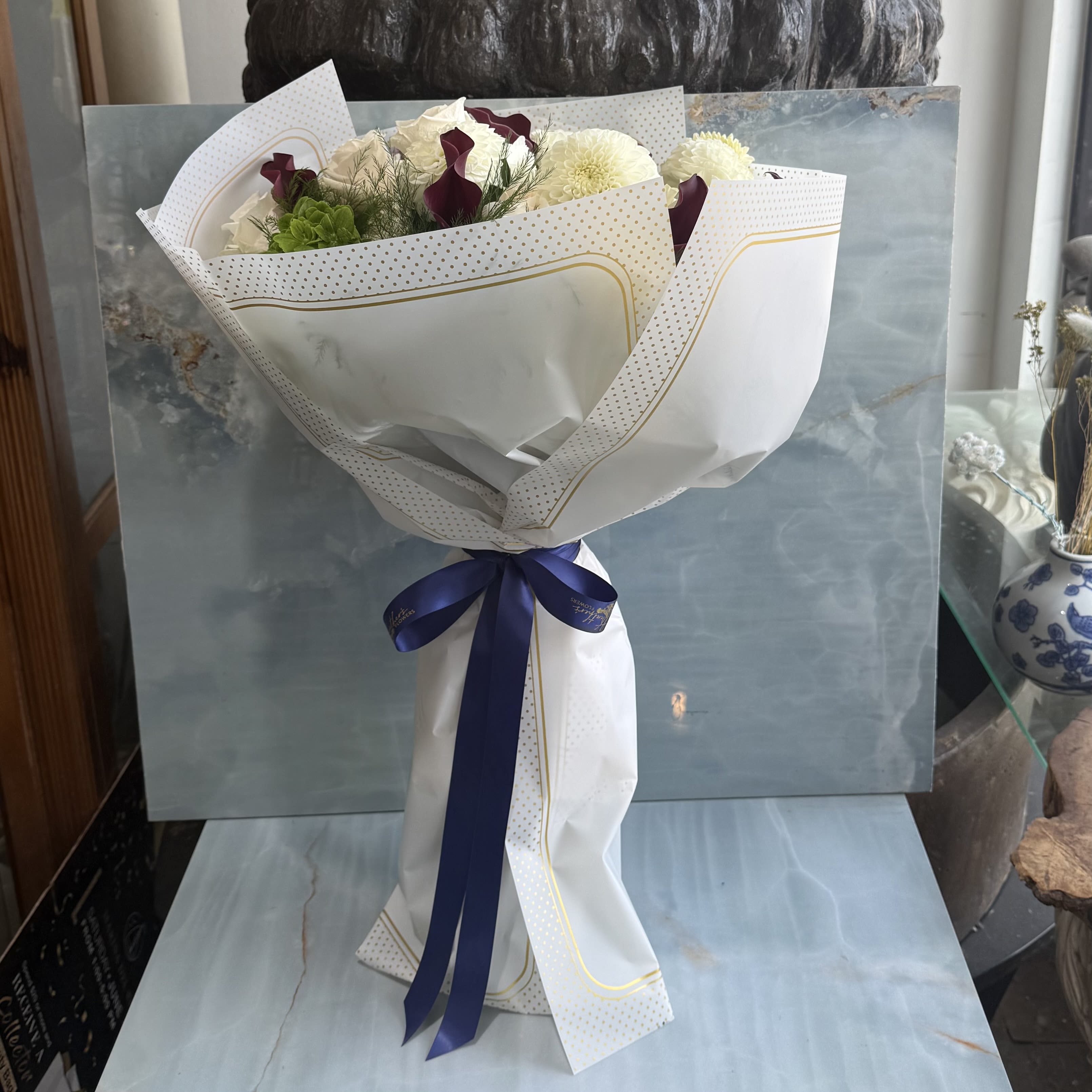 The Regal Edit  - Where timeless whites meet rich, moody tones. This hand-wrapped bouquet blends soft texture with striking contrast, finished with a tailored navy ribbon for a look that feels both modern and classic. Creamy white roses and white dahlias forms the heart of the bouquet, giving it that soft romantic feel.