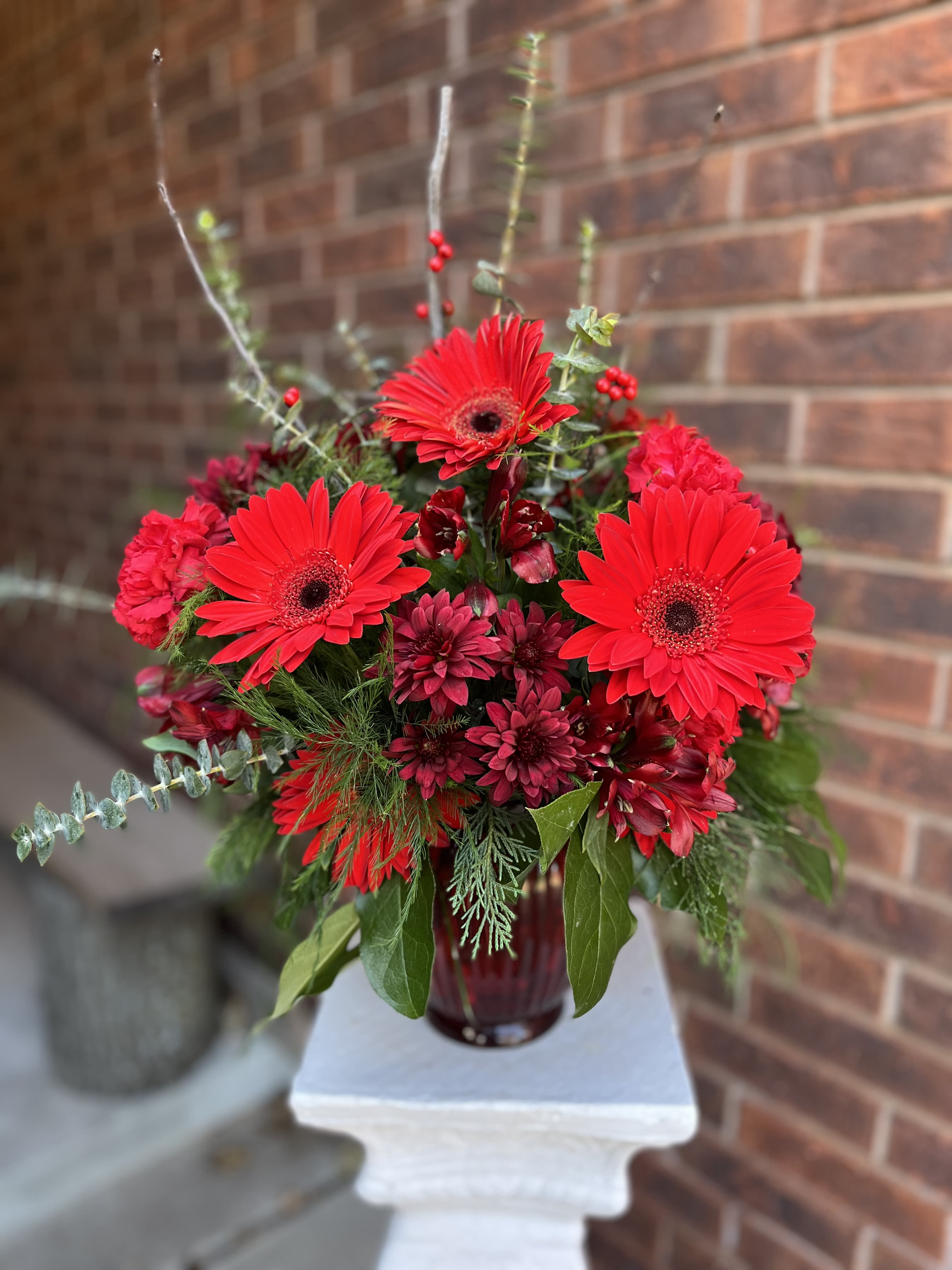 Lady in Red - Bright red gerbera daisies take center stage in this celebratory flower arrangement, making it an ideal choice for a birthday bouquet, anniversary roses alternative, or a festive holiday gift. The combination of vivid reds and deep wine tones gives the piece a bold, dramatic presence that will make the person you're thinking of feel truly celebrated.  This compact, rounded design pairs bright gerbera daisies with deep burgundy chrysanthemums and airy branching accents for height and movement, all set in a dark red glass vase. The mix of red, pink, and green hues provides contrast and depth-vibrant petals against rich, velvety tones-while the glass vase adds a polished, seasonal touch.  Perfect for hearth-side displays, dinner table centerpieces, or to send as a cheerful get-well or congratulations arrangement, Lady in Red reads as both joyful and refined. Arrives ready to enjoy in its vase; refresh water every 2-3 days and trim stems slightly to extend bloom life.